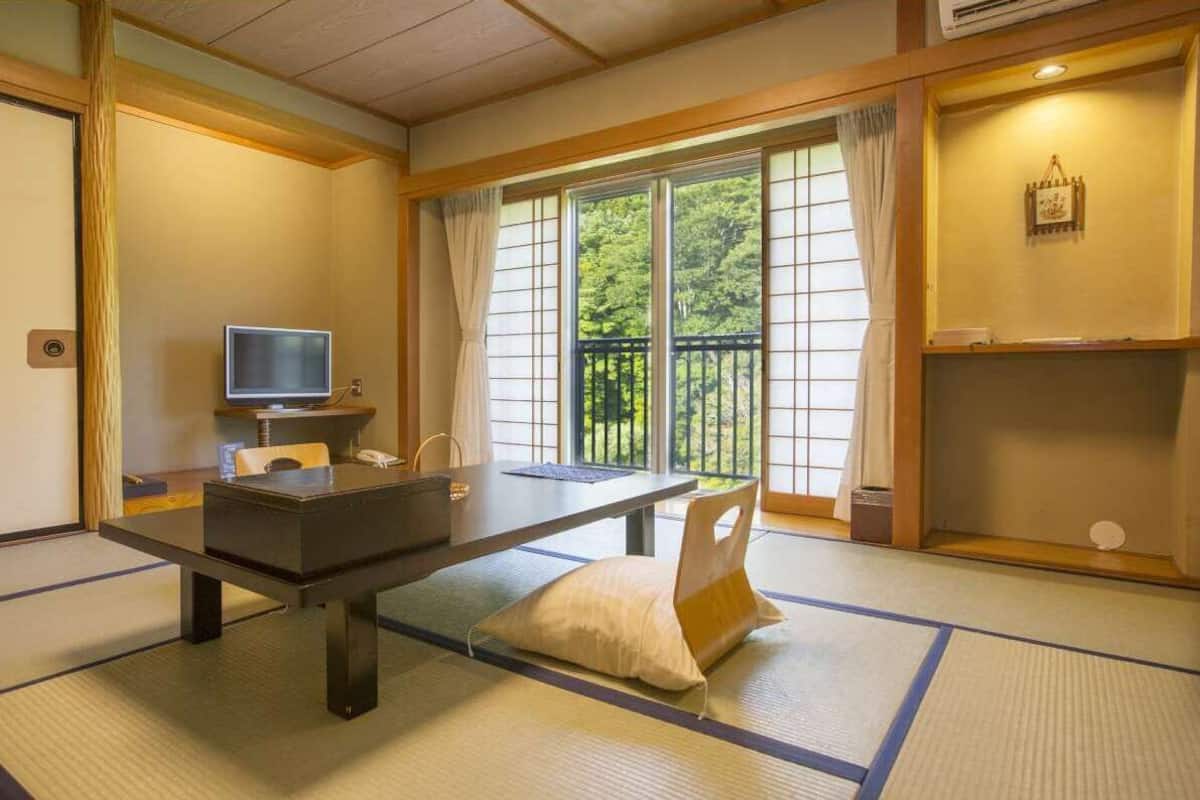 Ein traditioneller japanischer Raum mit Tatami-Boden, einem niedrigen Tisch, einem Fernseher und einem Balkon mit Blick auf Grünflächen.