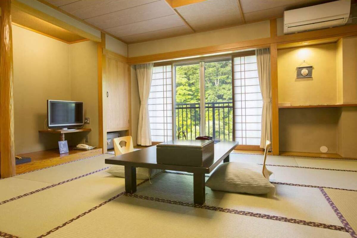 Ein traditioneller japanischer Raum mit Tatami-Boden, einem niedrigen Tisch, einem Fernseher und einem Balkon mit Blick auf Grünflächen.