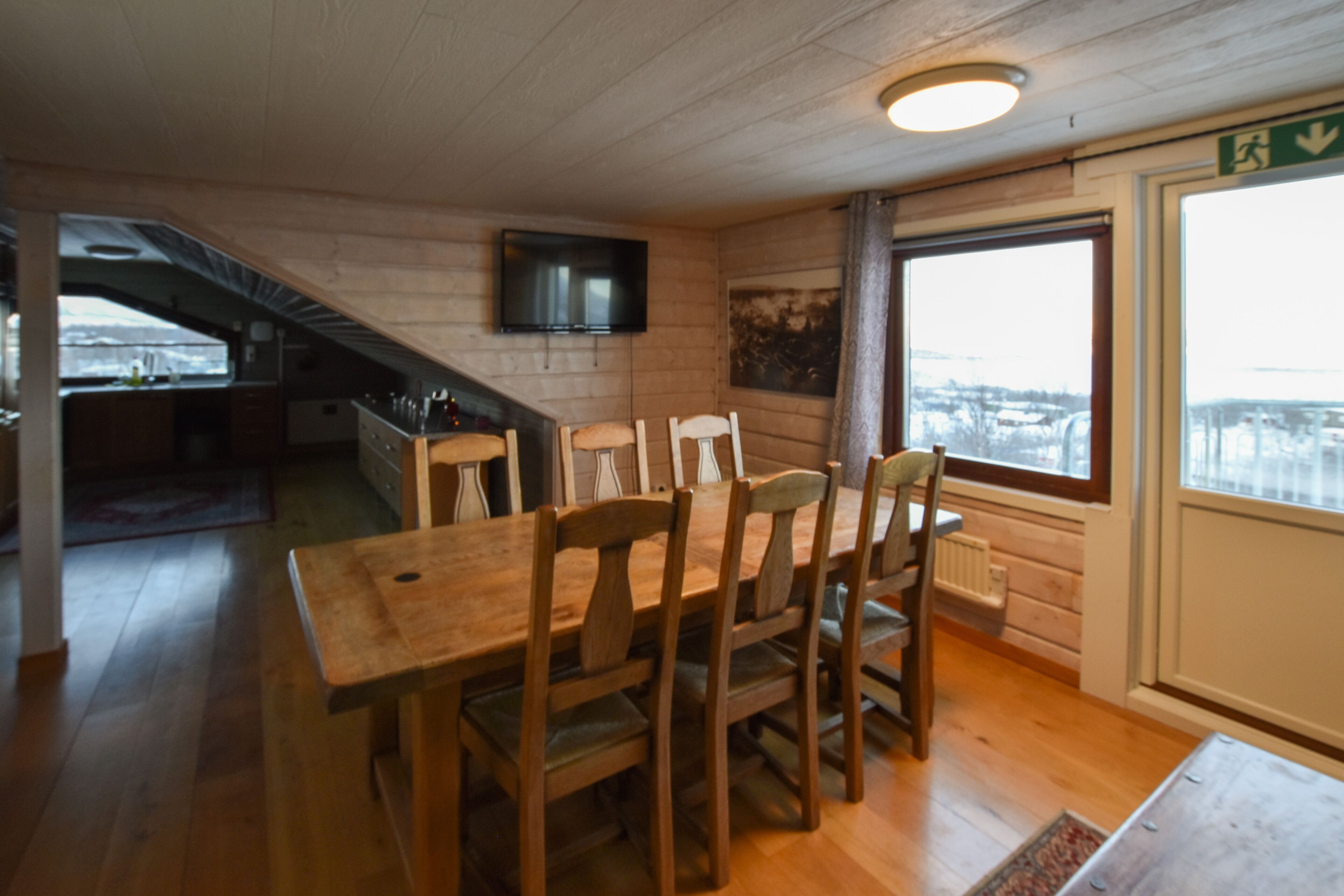 Una mesa de comedor de madera con sillas, un televisor montado en la pared y una ventana con vistas al exterior.