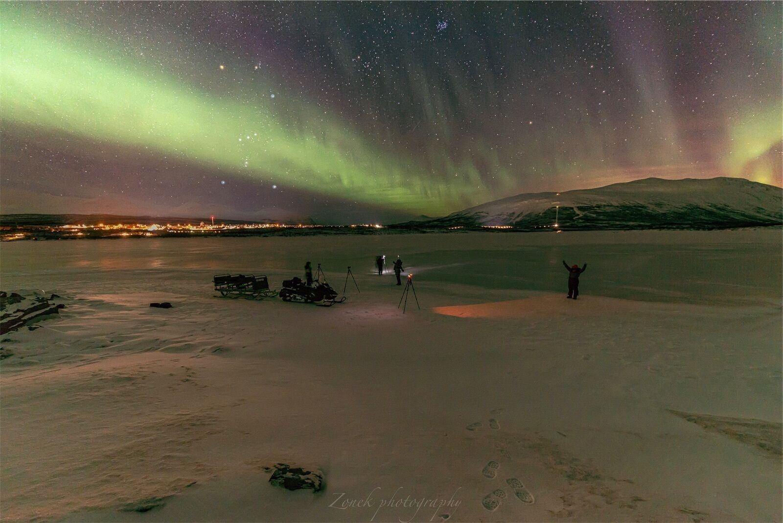 A snowy landscape with a person standing on a sled, a snowmobile, and a tripod with lights.