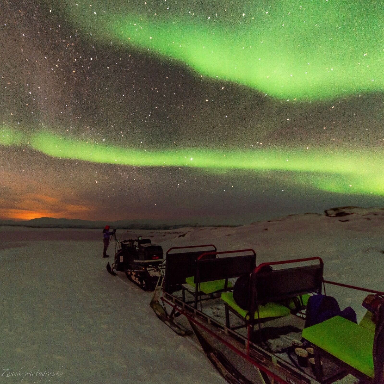 A snow-covered landscape with a sled pulled by a dog team, under a night sky with visible stars and the aurora borealis.