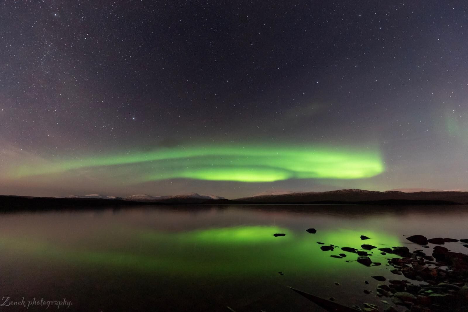 A night sky with stars and the Northern Lights reflected on a calm lake.