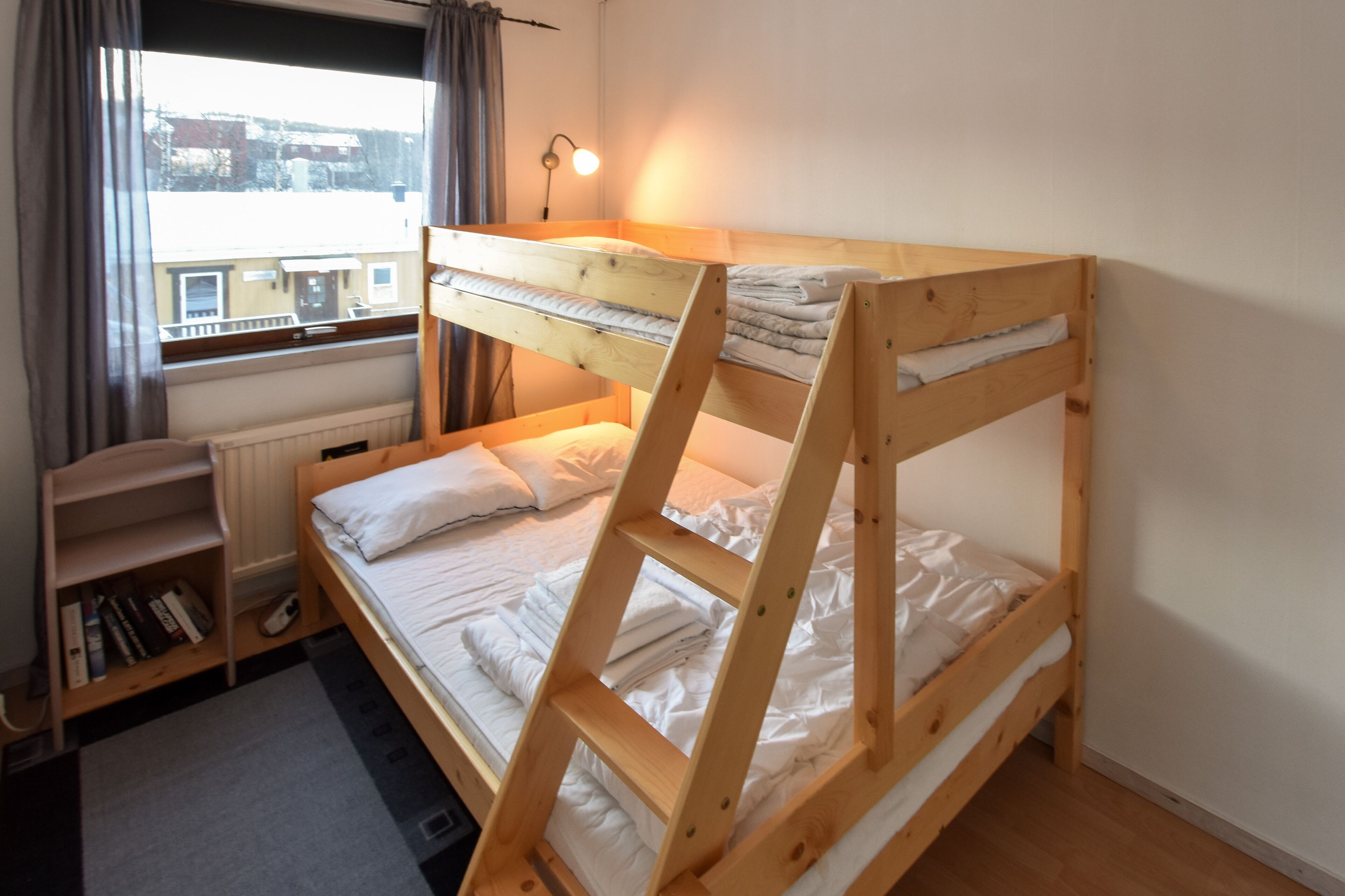 A bunk bed with a wooden frame, a bedside table with a lamp, a radiator, and a window with curtains.