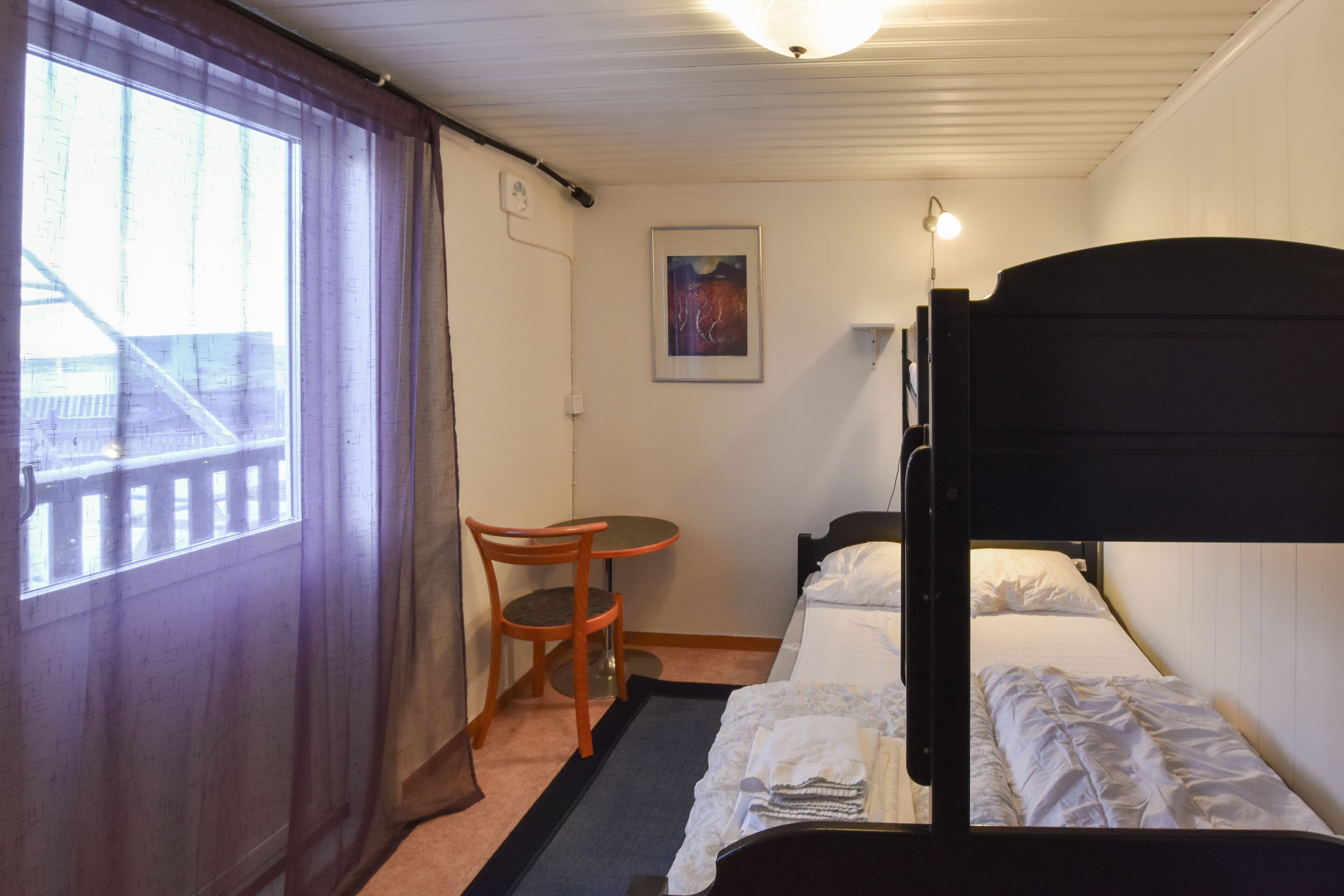 A small, single-bed room with a wooden bunk bed, a small table with a chair, a window with curtains, and a framed picture on the wall.