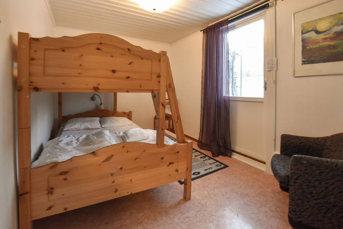 Ein hölzernes Etagenbett mit einer Leiter, ein Einzelbett und ein Stuhl in einem Raum mit Fenster und einem Bild an der Wand.