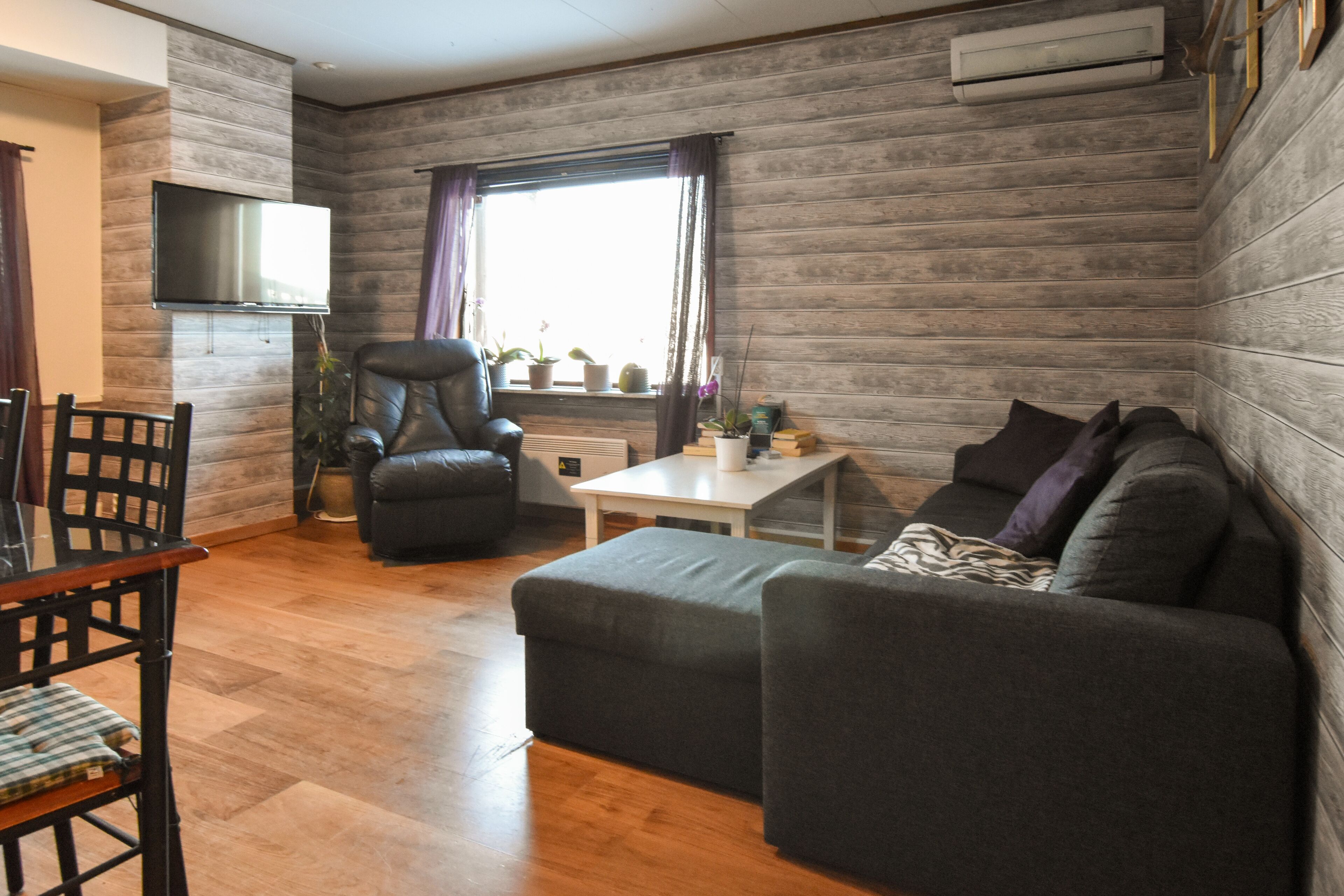 A cozy living room with a flat-screen TV mounted on the wall, a comfortable sofa, a dining table, and a window with curtains.
