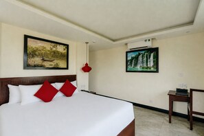 Quarto de hotel com uma cama grande, duas pinturas nas paredes, uma luminária pendente vermelha e uma mesinha com um controle remoto.