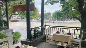 An outdoor dining area with tables set for two, featuring a view of a river and a cityscape in the background. - River Suites Hoi An Hotel (Da Nang)