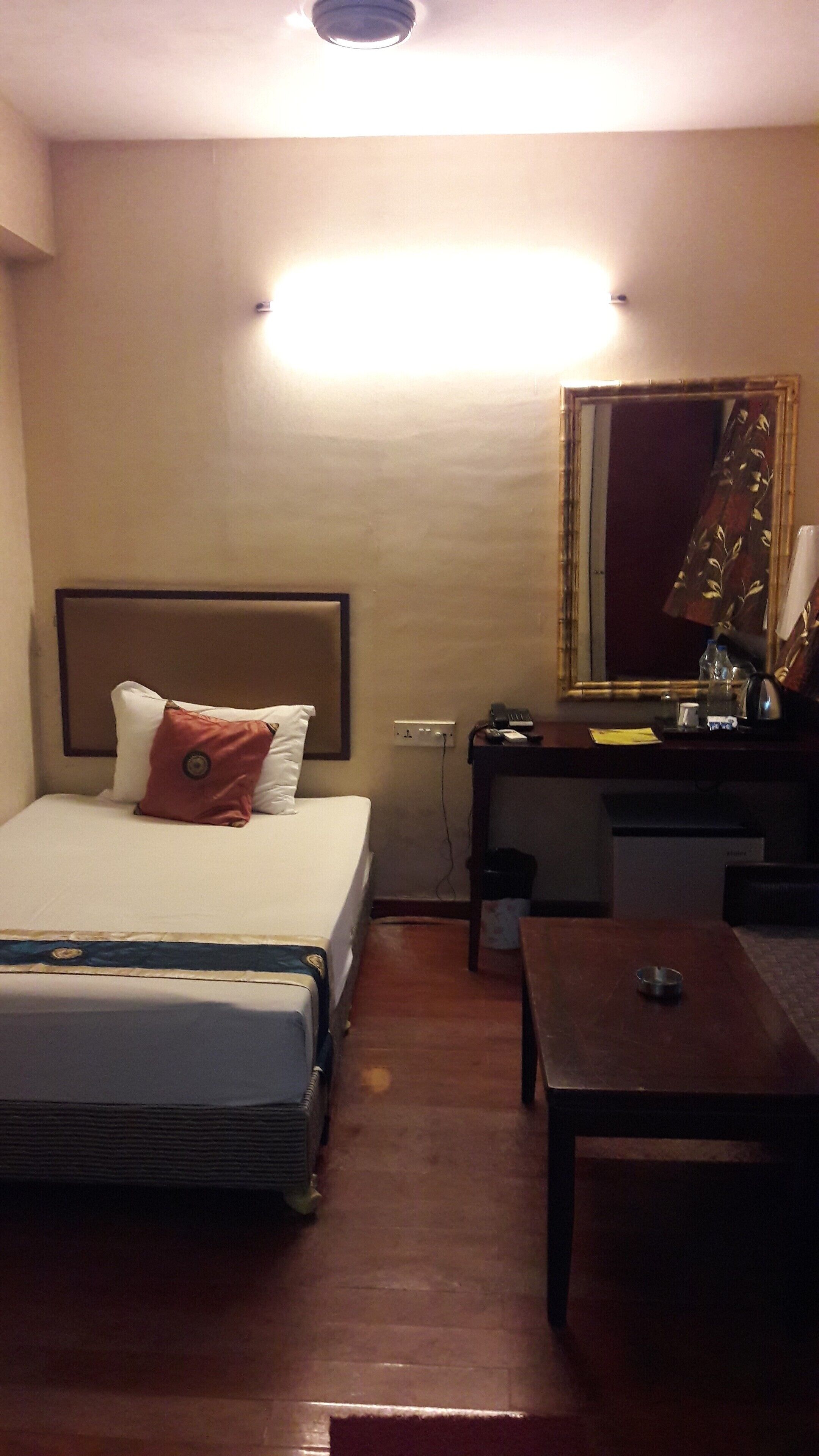 A hotel room with a bed, bedside table, desk, mirror, and a small table with a cup.