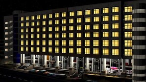 Un hotel moderno con una fila de ventanas uniformemente iluminadas, una entrada central con toldo y un área de estacionamiento al frente.