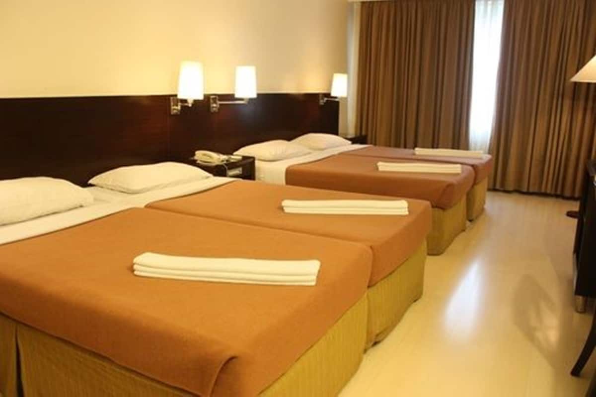 Een hotelkamer met vier eenpersoonsbedden, elk voorzien van een bruine sprei en witte kussens. Twee wandlampen zitten op het hoofdeinde, en aan de rechterkant is een raam met gordijnen.
