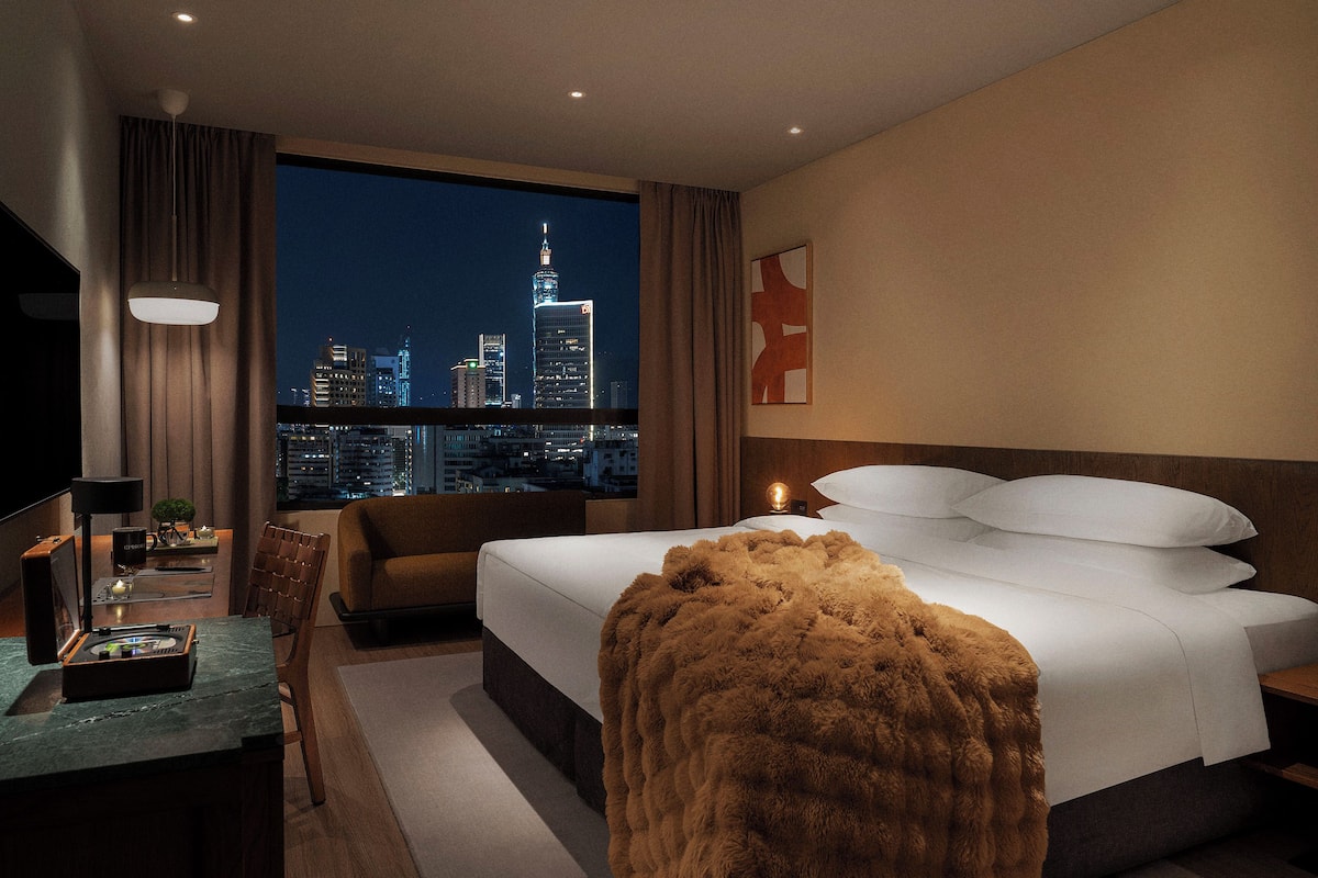 A modern hotel room with a large bed, a desk with a lamp, a chair, and a view of the city skyline at night.