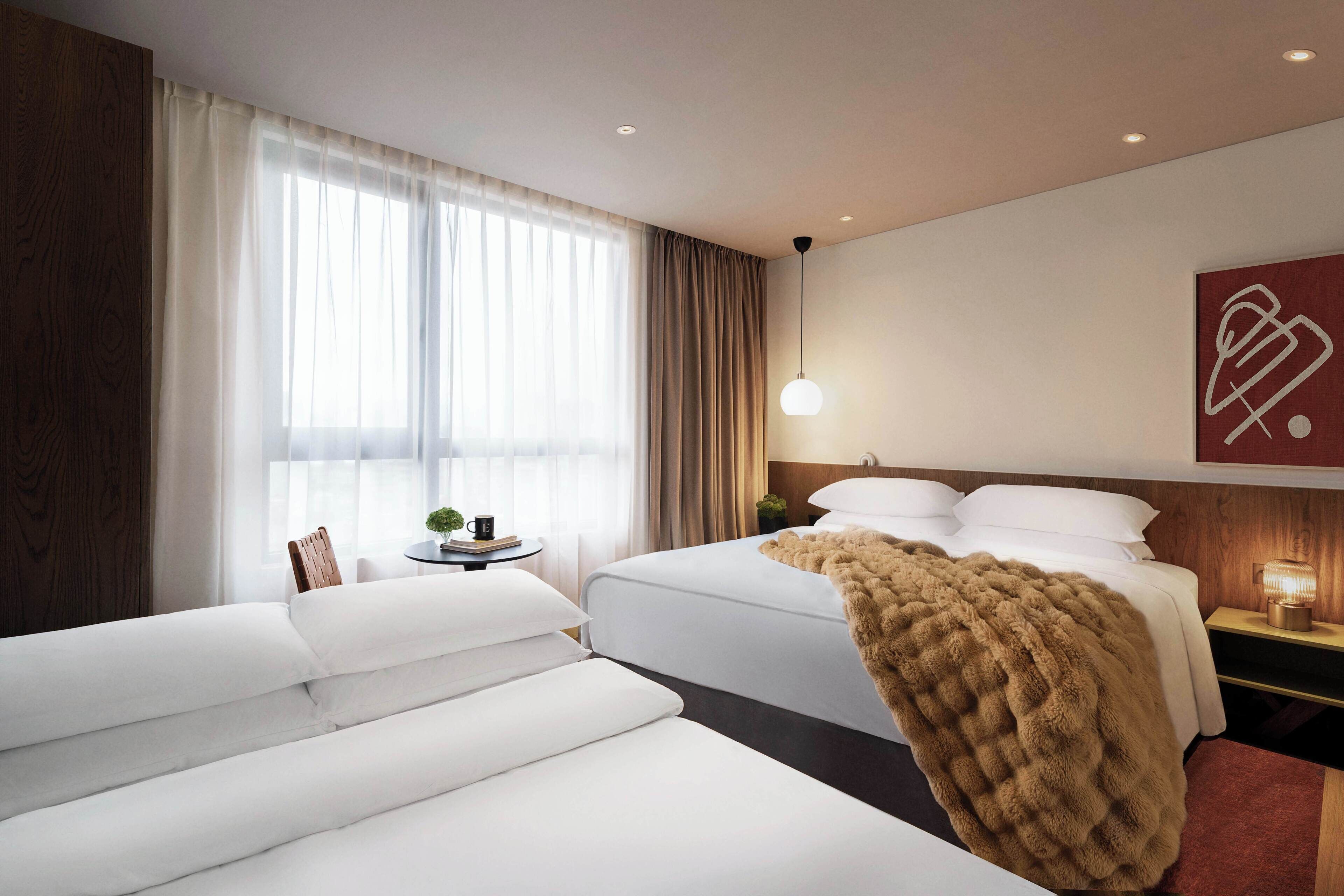Une chambre d’hôtel avec deux lits, un canapé, une table de chevet avec une lampe, une fenêtre avec des rideaux et une œuvre d’art murale.