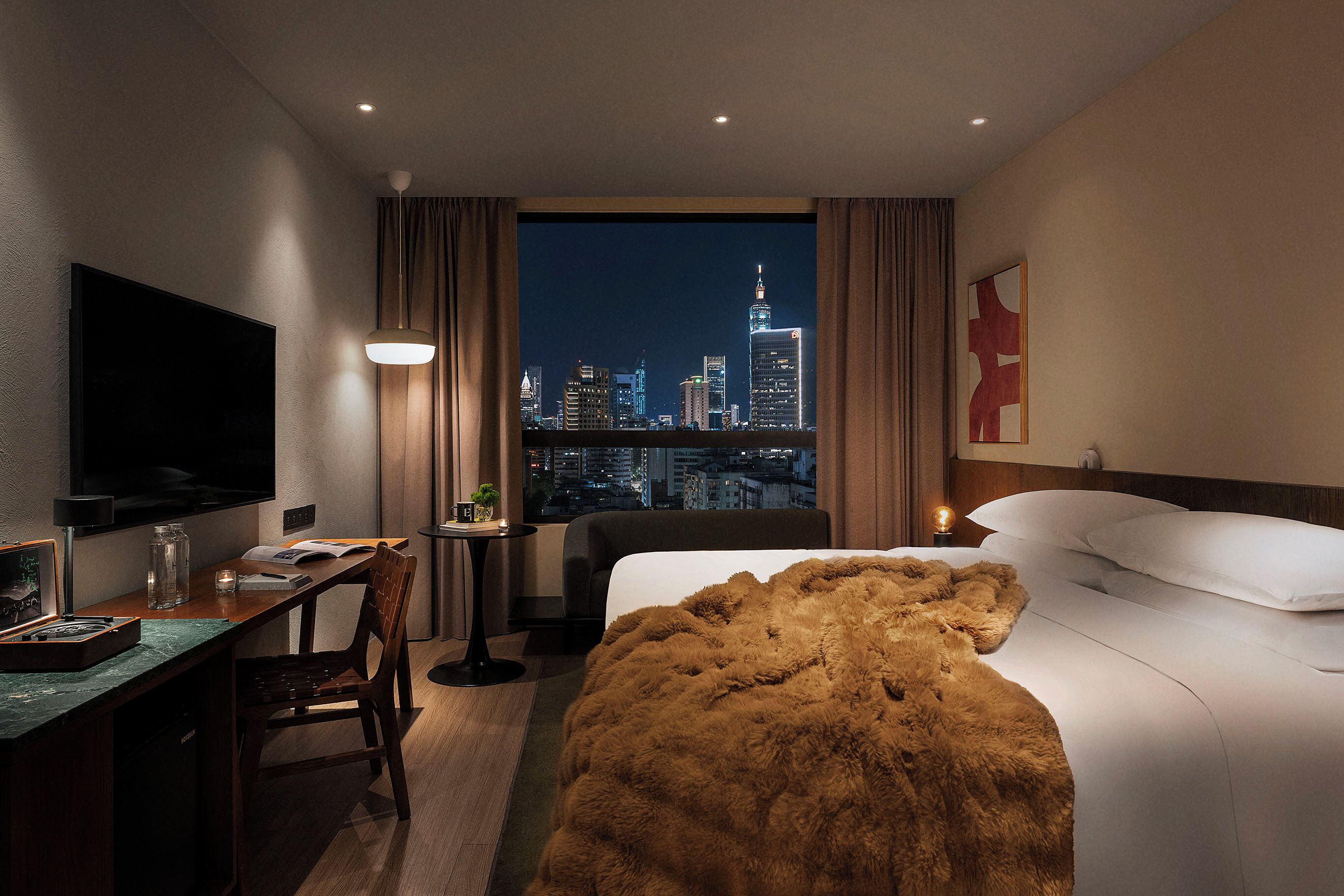 A hotel room with a bed, a desk, a chair, a TV, and a view of a city skyline at night.