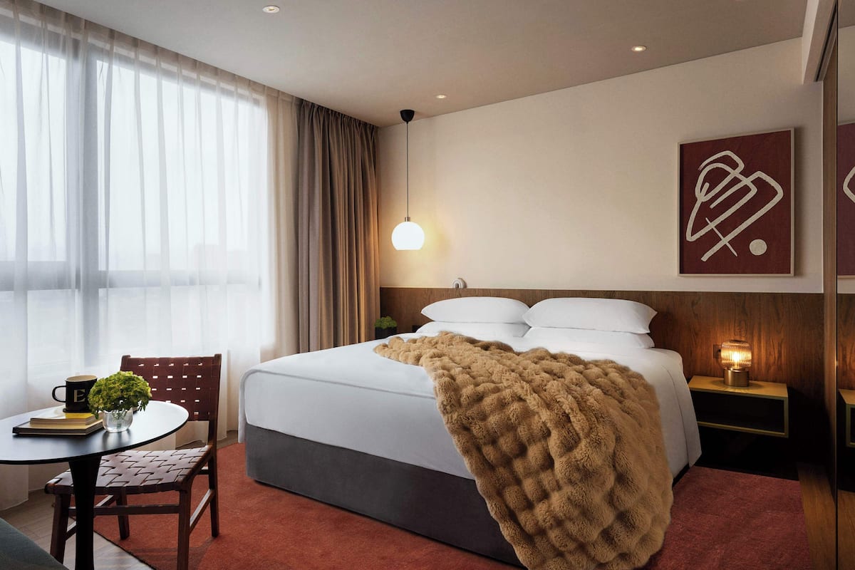 A hotel room with a bed, a bedside table with a lamp, a chair, a small table with a plant, and a window with curtains.