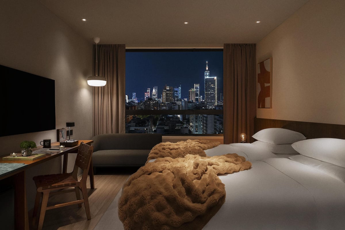 A hotel room with a city view, a desk, a chair, a sofa, and a bed with a fluffy blanket.