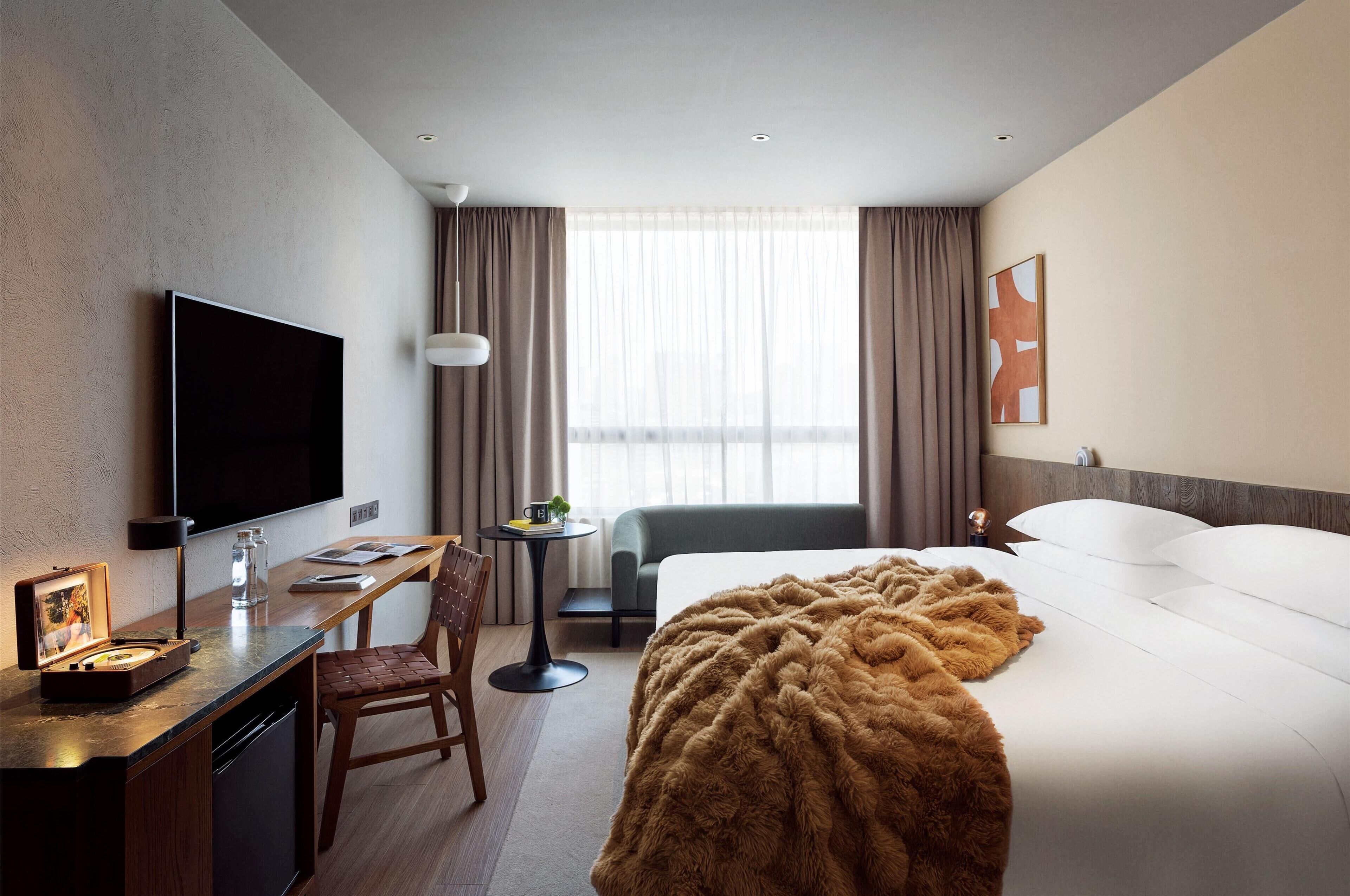 A hotel room with a bed, a desk with a flat-screen TV, a chair, a small table with a lamp, and a window with curtains.