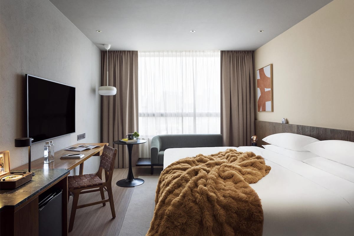 A hotel room with a bed, a desk with a TV, a chair, a table with a lamp, and a window with curtains.