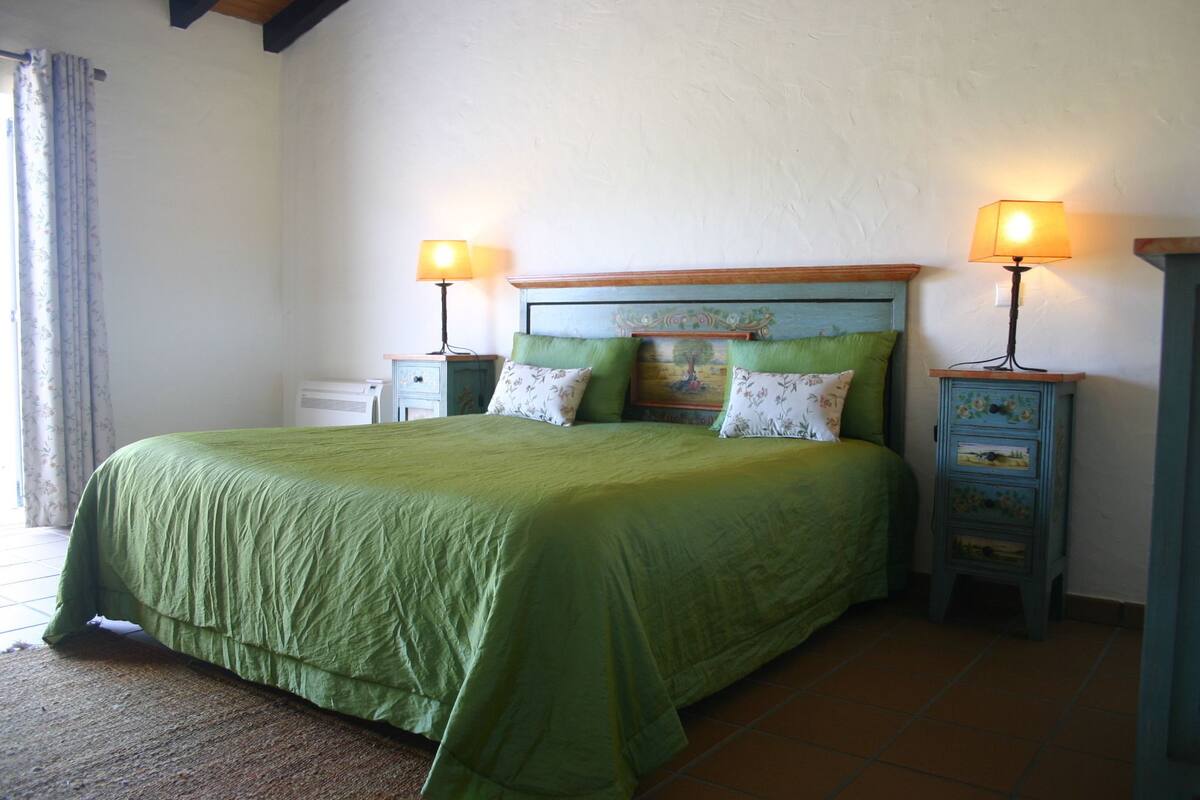 A bed with a green bedspread, two bedside tables with lamps, and a small dresser with floral patterns.