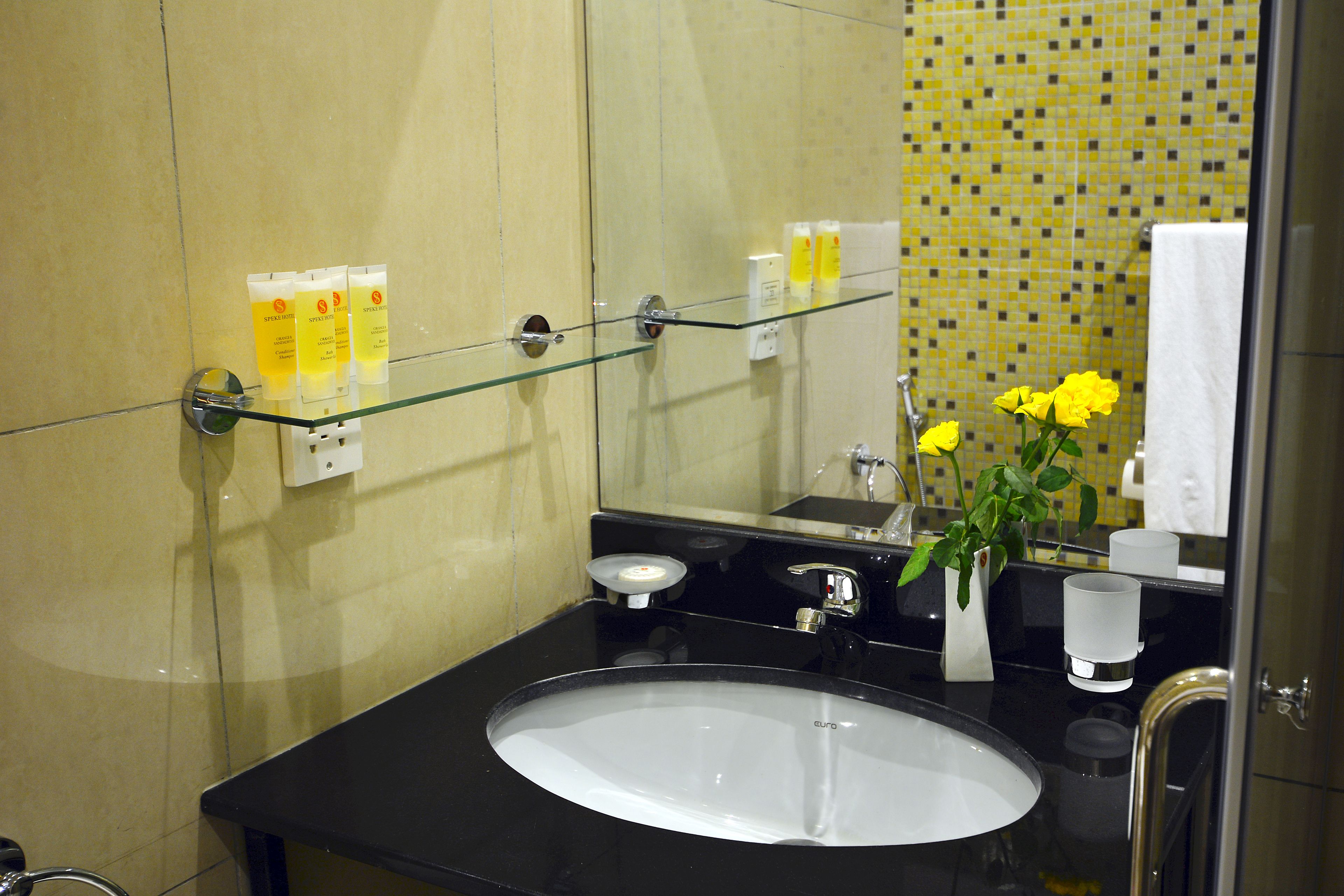 Un baño con un lavabo negro, repisas de vidrio, flores amarillas y una pared con azulejos amarillos.