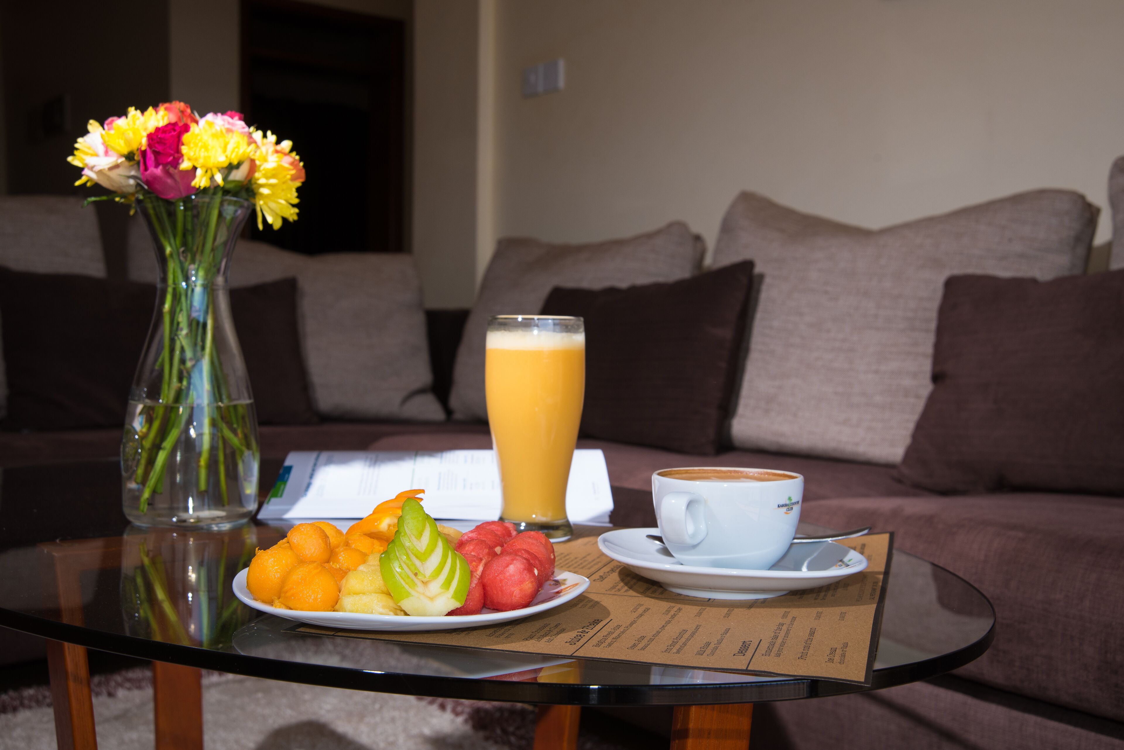 Une table mise pour le petit-déjeuner, avec une assiette de fruits, un verre de jus d’orange, une tasse de café et un vase de fleurs posé sur une table en verre.