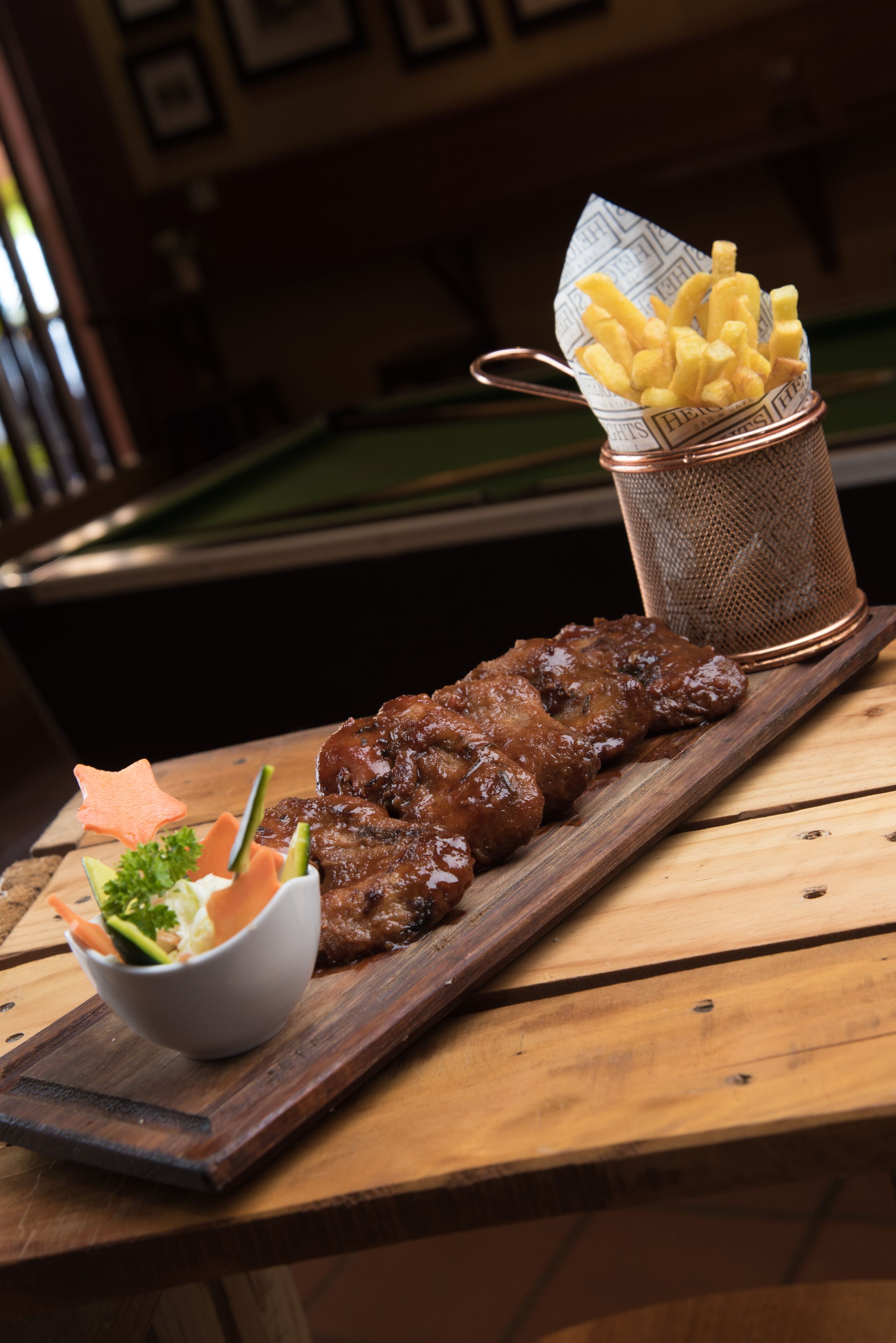 Una mesa de madera con un plato de carne glaseada, un tazón pequeño de ensalada y una cesta de papas fritas.