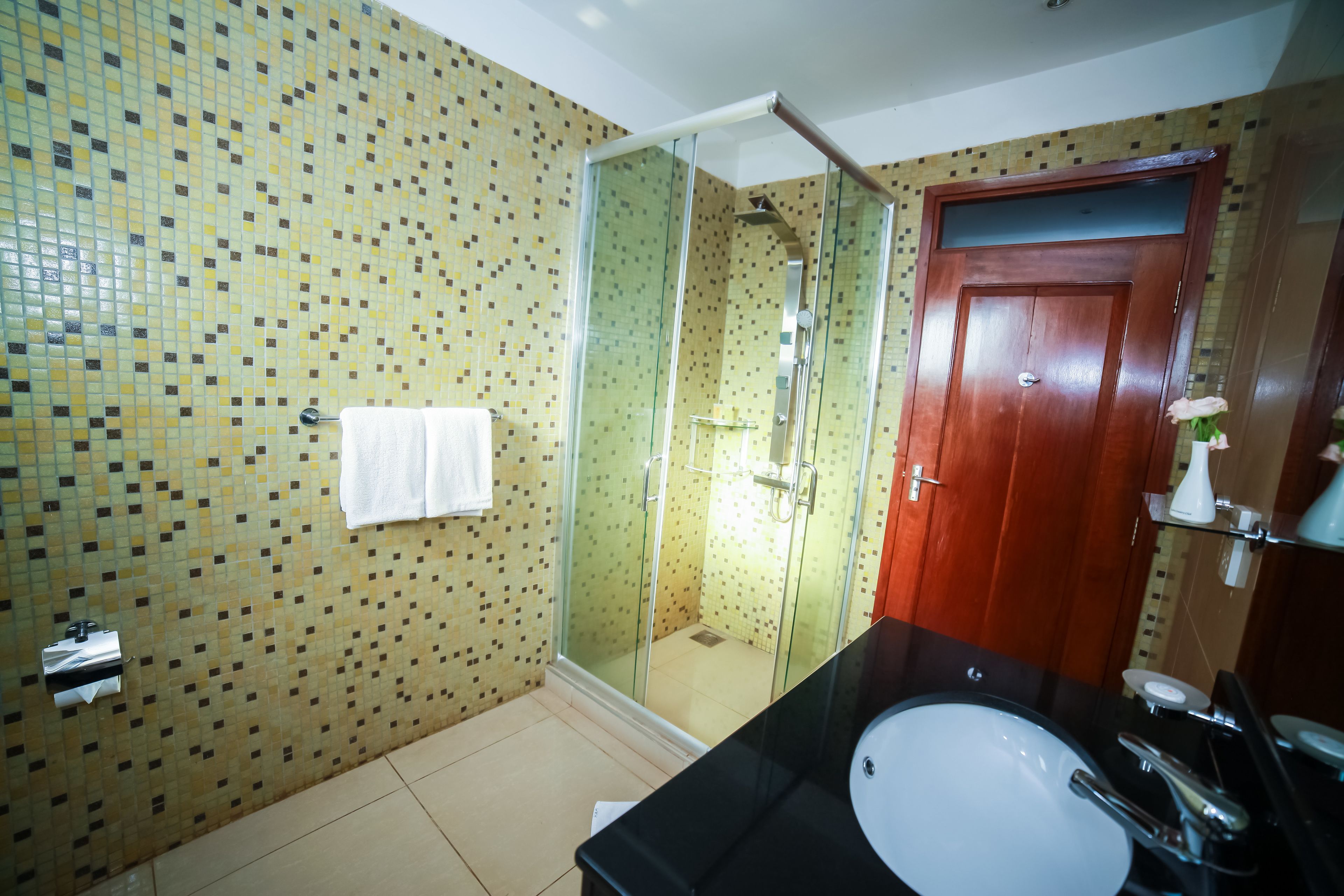 Un baño con pared de azulejos, cabina de ducha de vidrio, lavamanos negro y puerta de madera.
