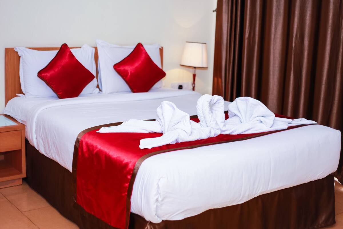 A neatly made bed with red and white bedding, a wooden headboard, a nightstand with a lamp, and a brown curtain.