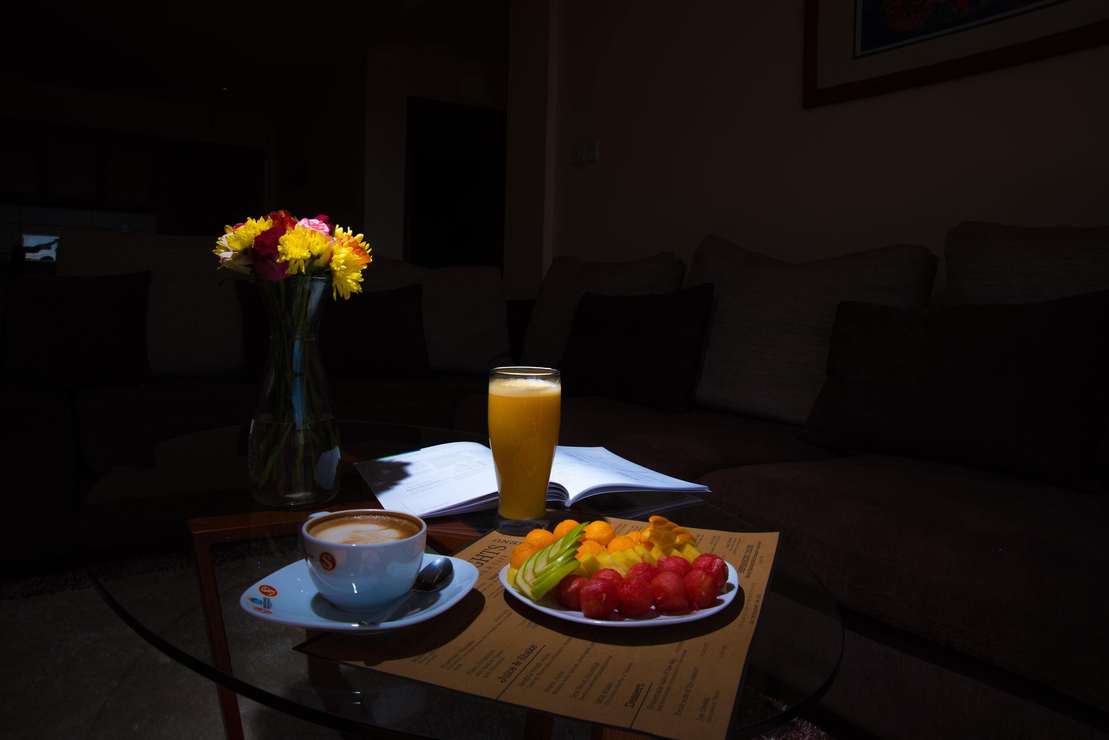 Une table avec une tasse de café, un verre de jus d’orange, une assiette de fruits et un vase de fleurs.