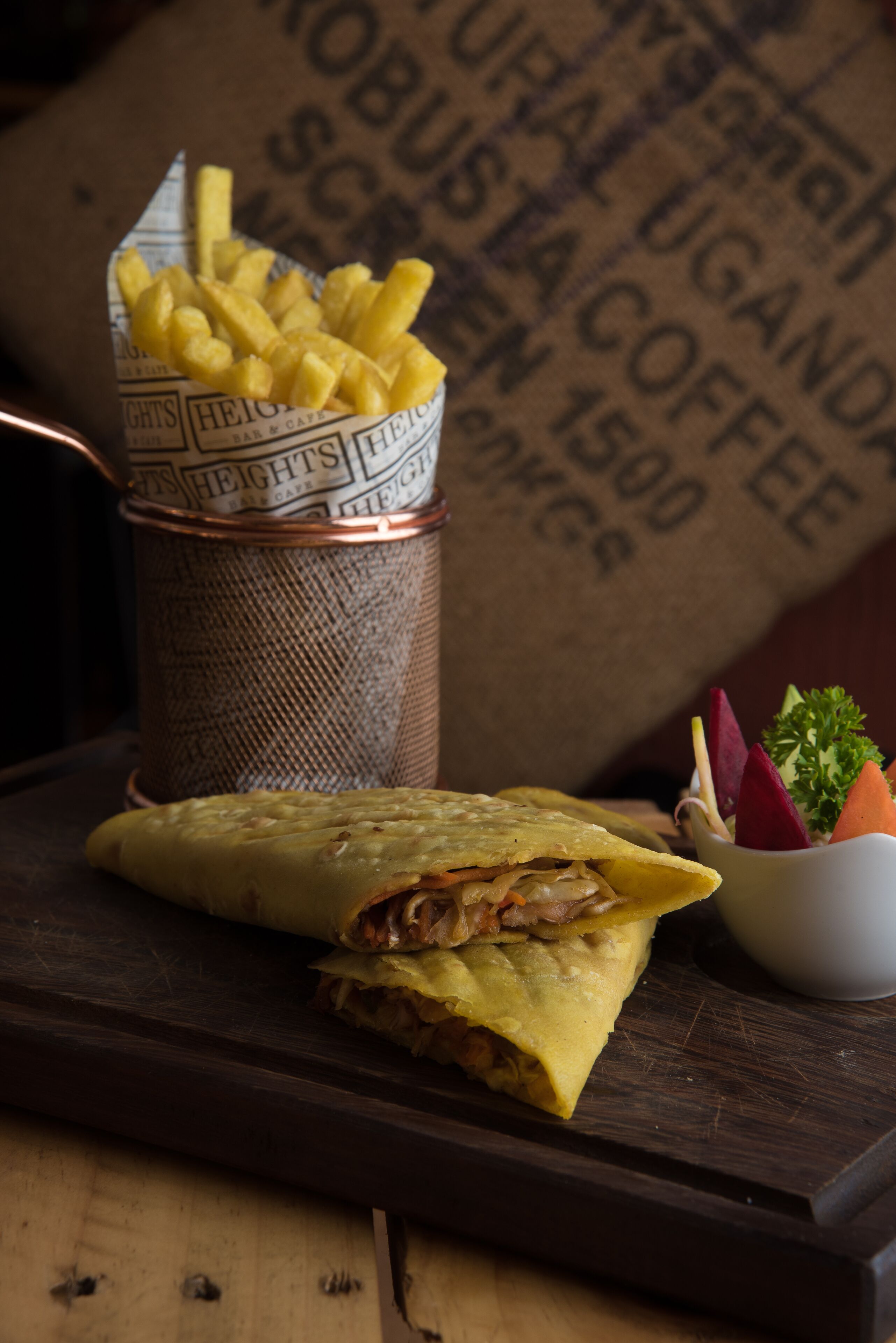A sandwich on a wooden board, a cup of fries, and a small bowl of vegetables.