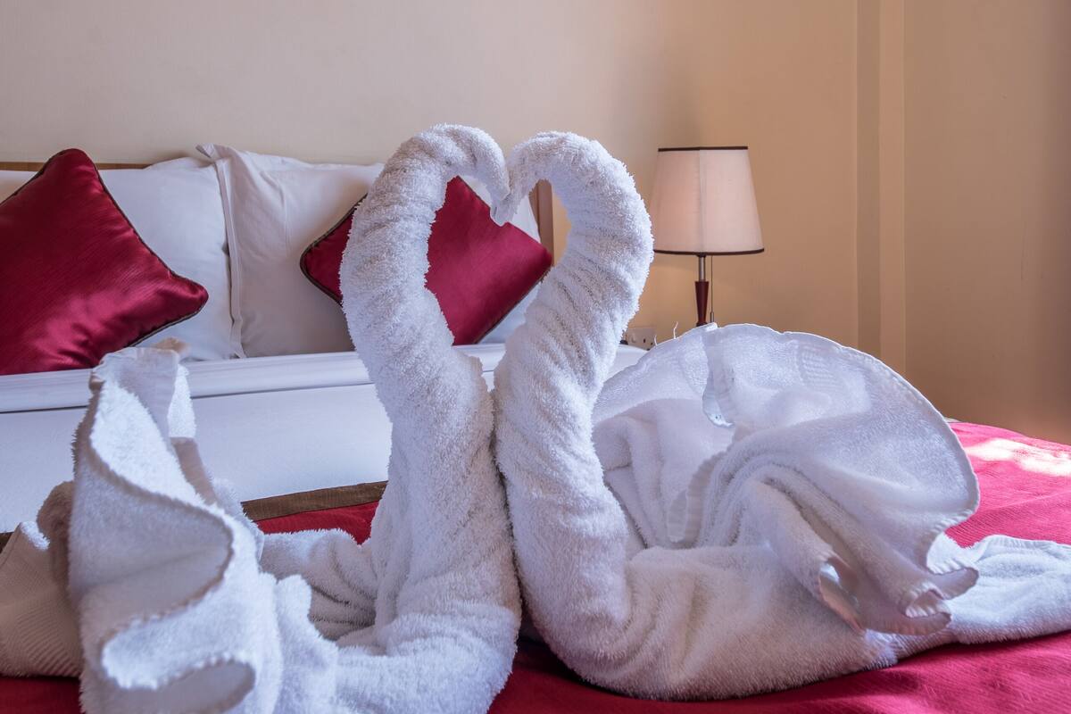 A hotel room with a bed, red and white pillows, a lamp, and two towels arranged in a heart shape.
