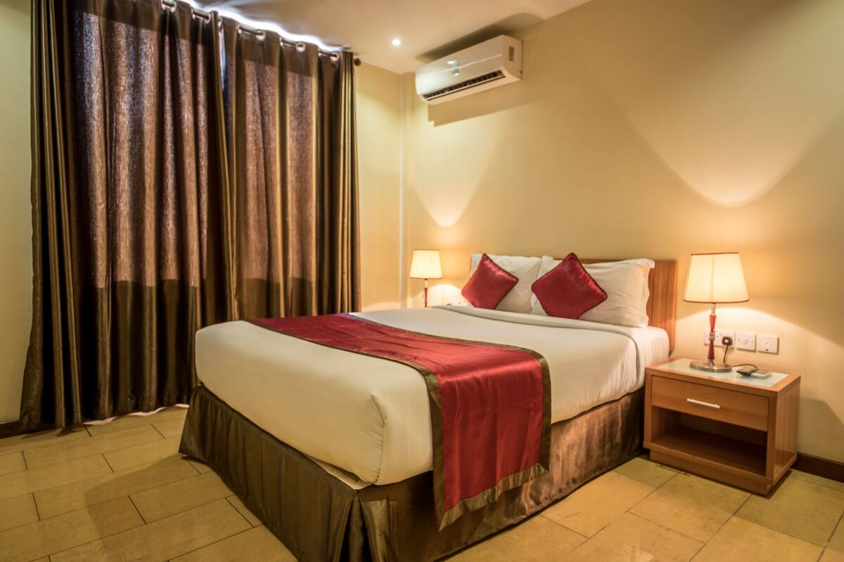 A hotel room with a bed, curtains, bedside lamps, a nightstand, and an air conditioning unit.