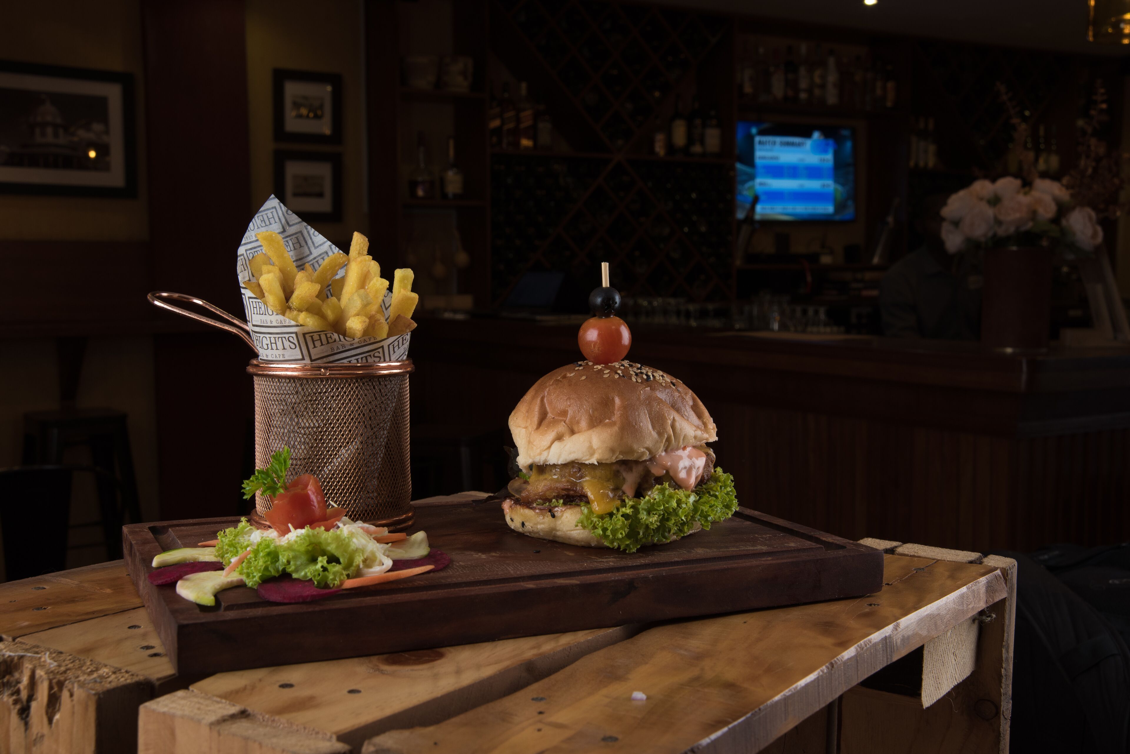 A burger with a cherry on top, a basket of fries, and a salad on a wooden table in a bar setting.