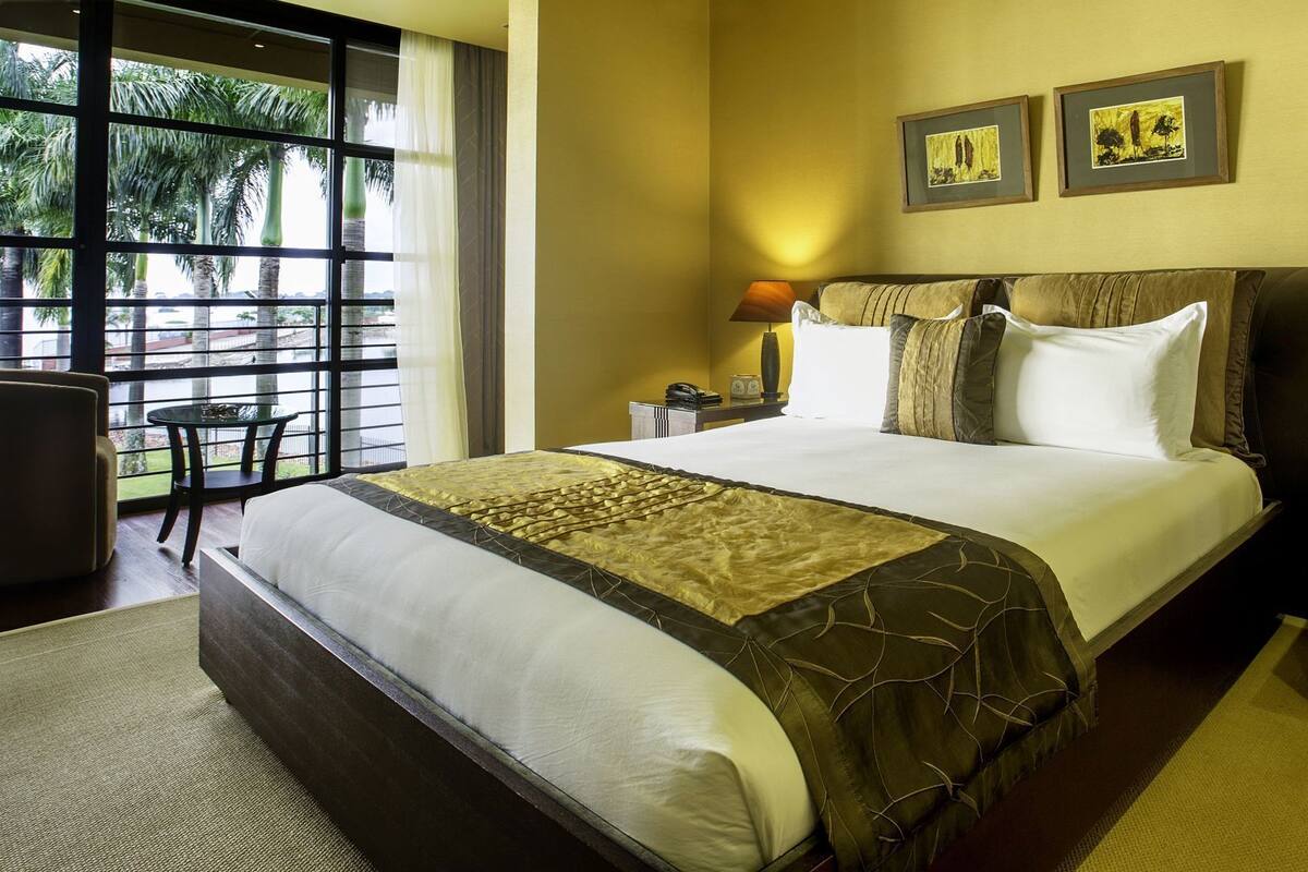 Habitación de hotel con una cama grande, dos lámparas de noche, una mesita de noche, una silla y vistas a palmeras a través de la ventana.