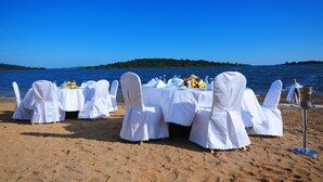 Montaje para cenar en la playa con manteles blancos y sillas, un cubo metálico y un arreglo floral.