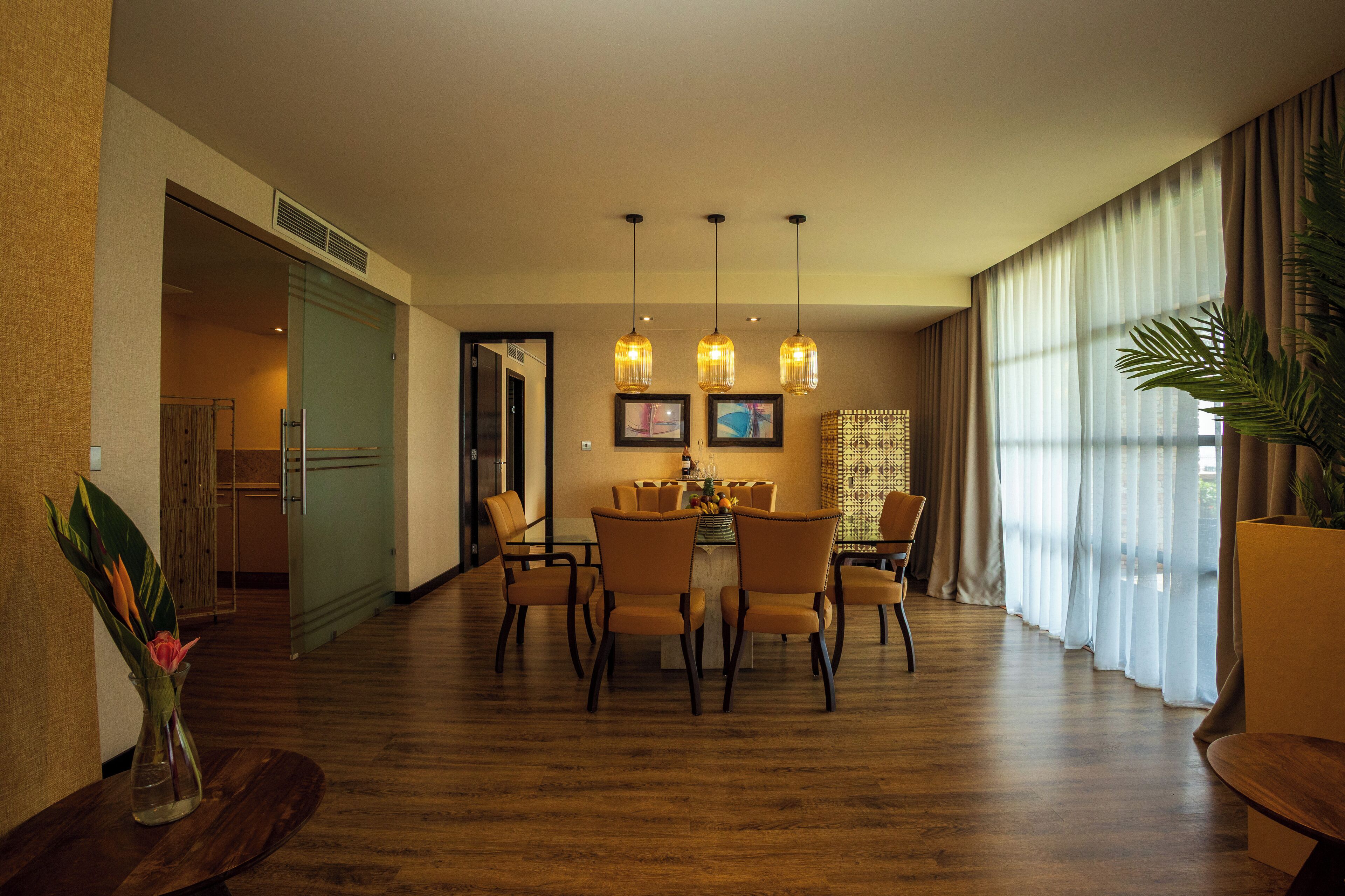 A hotel dining area with a wooden floor, a round table with chairs, pendant lighting, and a large window with sheer curtains.
