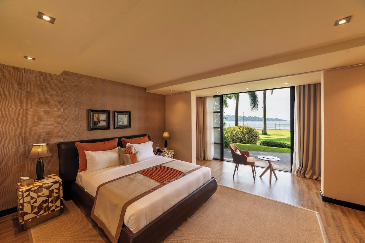 Habitación de hotel moderna con una cama grande, una mesita de noche, una lámpara, vistas al exterior y una pequeña mesa con sillas.