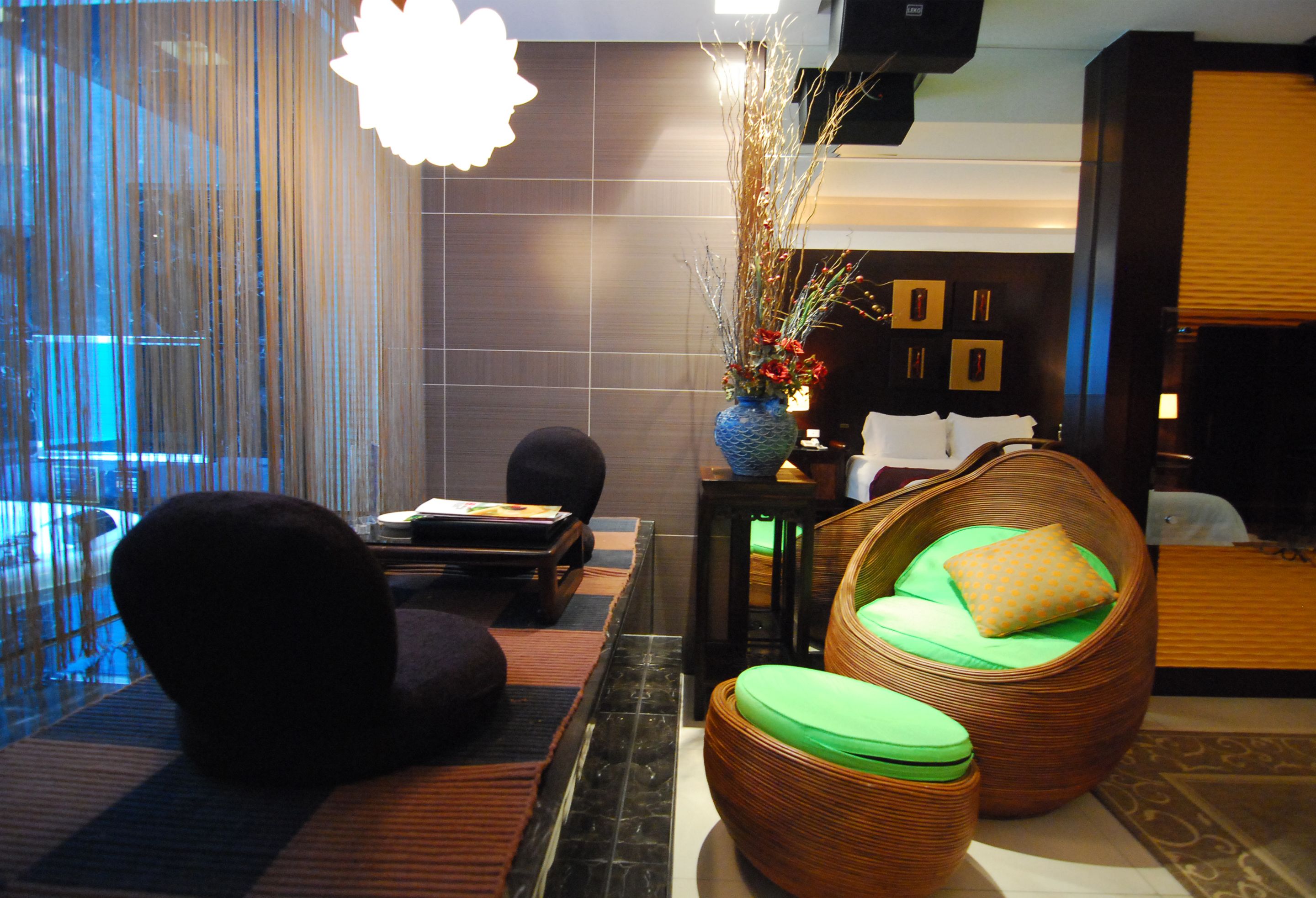 A modern lobby with a large window, a black sofa, a wicker chair with a green cushion, and a decorative vase with branches.