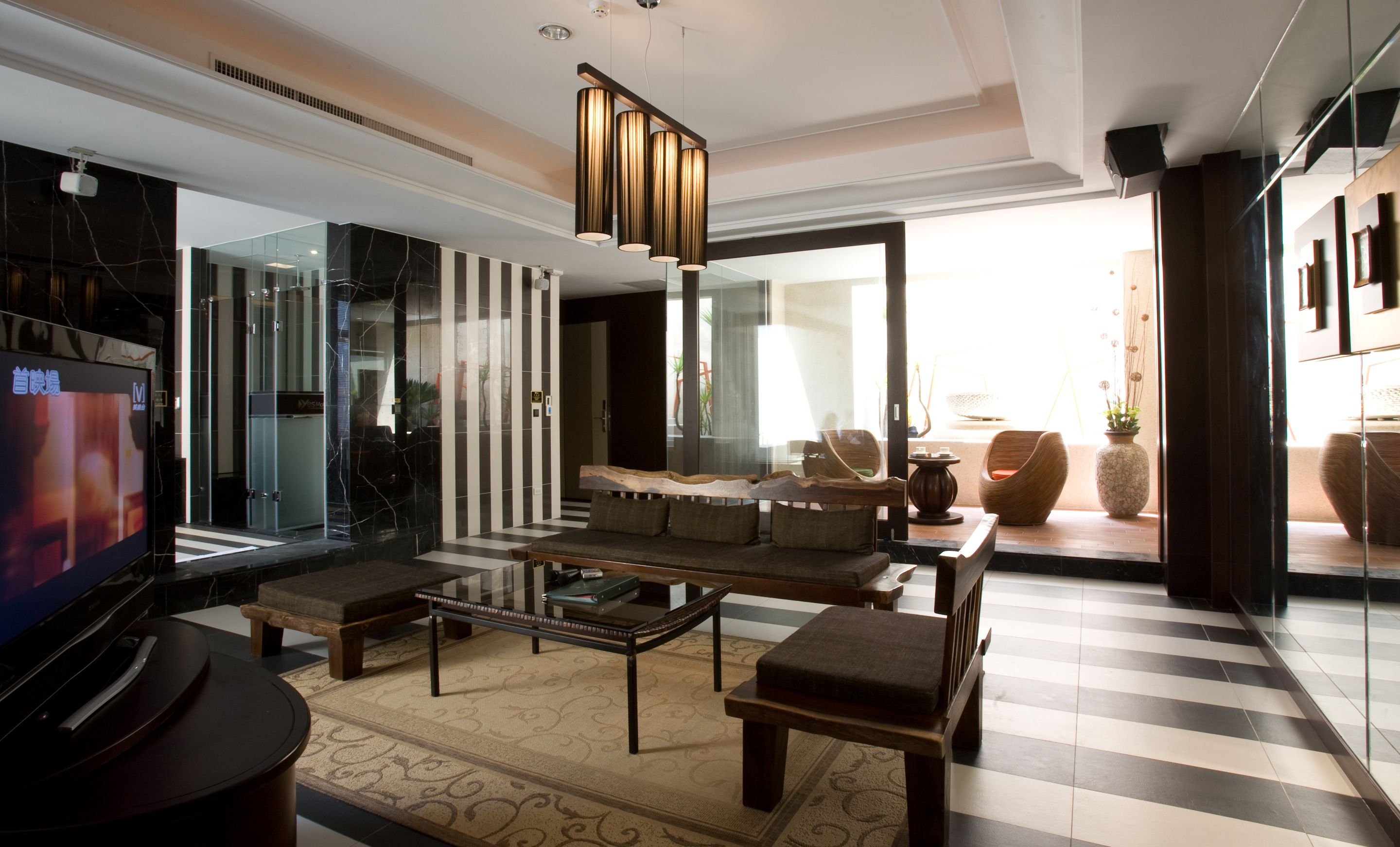 A modern living room with a large flat-screen TV, a wooden coffee table, and a sofa. There is a glass partition with a striped design and a large window providing natural light.