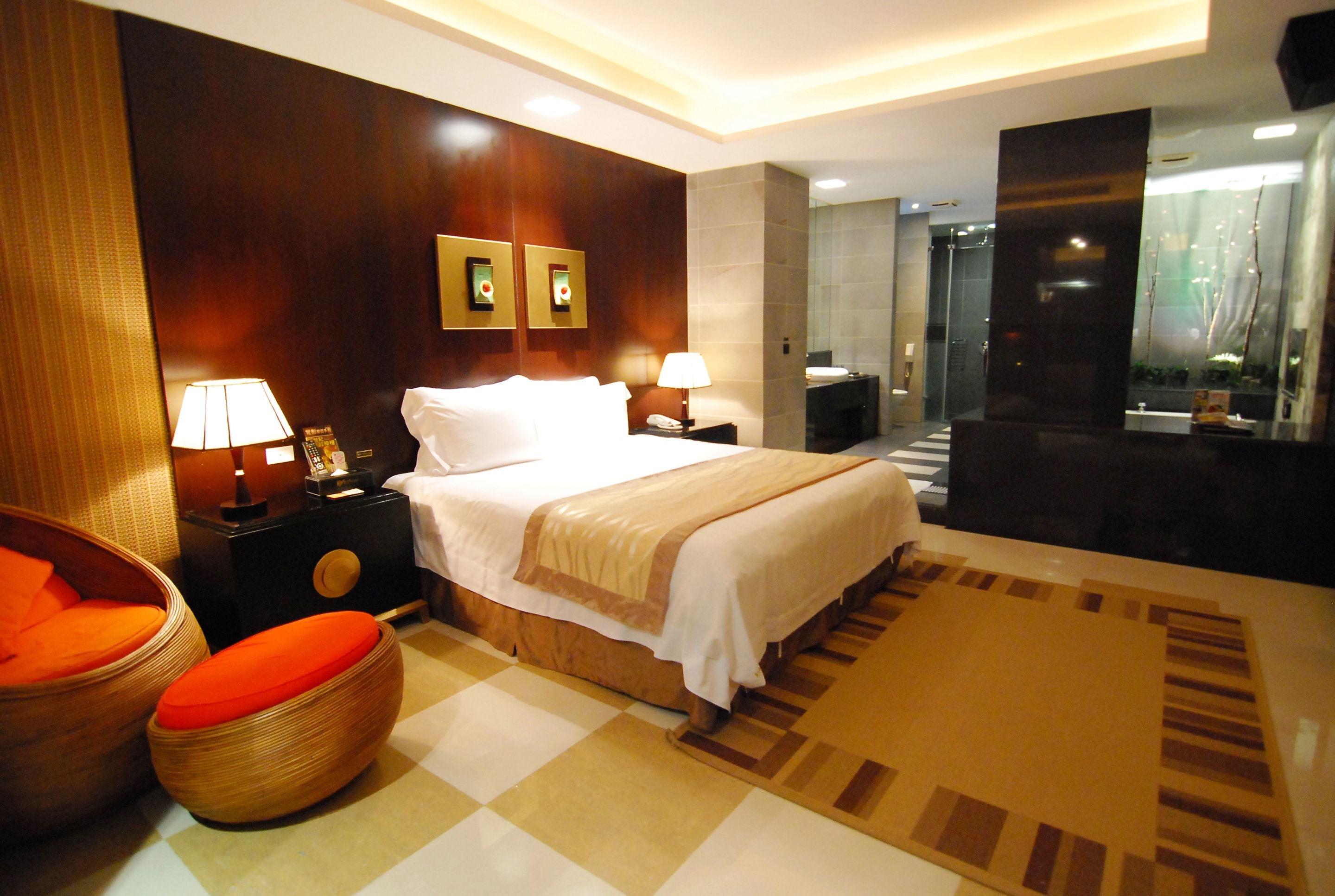 A modern hotel room with a large bed, two bedside lamps, a wooden nightstand, a wicker chair, and a bathroom visible through a glass door.