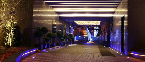 A hotel corridor with a checkered floor, illuminated by underglow lighting, and lined with potted plants and decorative lights.