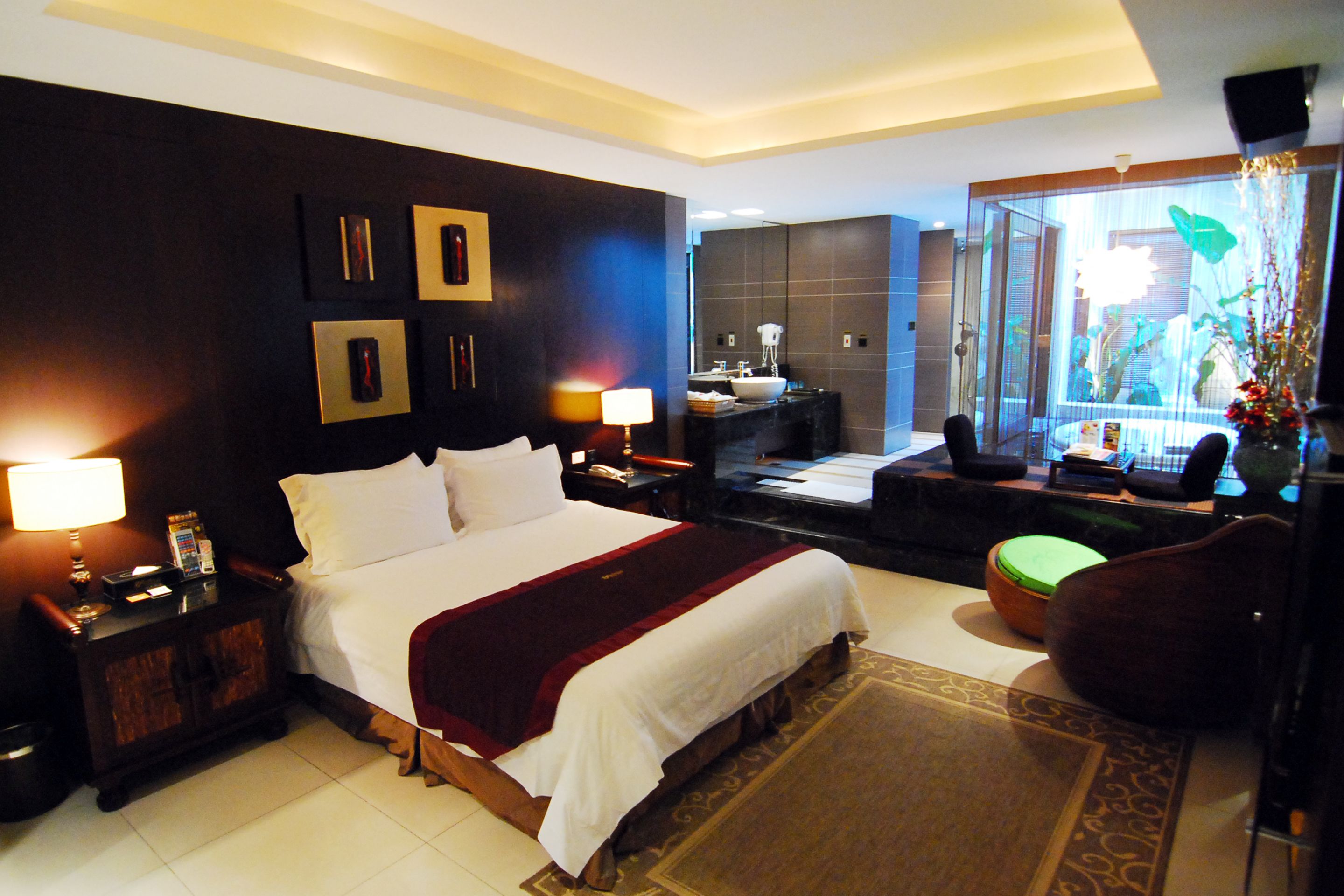 A modern hotel room with a large bed, a sitting area with a sofa and armchair, a small table, and a bathroom with a vanity and mirror.