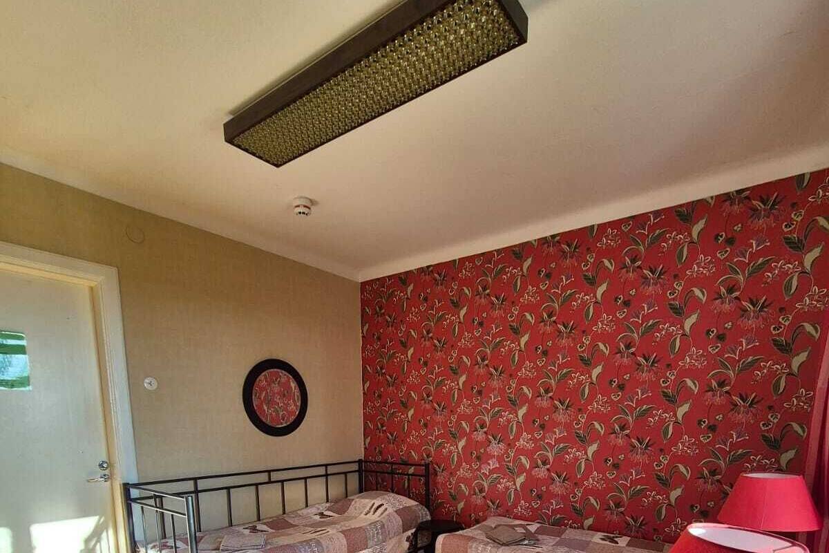 Une chambre avec deux lits simples, un papier peint à motifs floraux rouges, un plancher en bois, une lampe rouge et un luminaire au plafond.