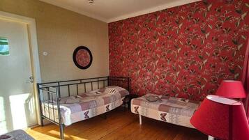 Una habitación con dos camas individuales, papel tapiz rojo con estampado floral, piso de madera, una lámpara roja y un accesorio de iluminación en el techo.