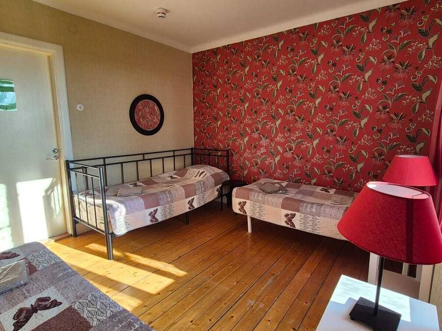 Una habitación con dos camas individuales, papel tapiz rojo con estampado floral, piso de madera, una lámpara roja y un accesorio de iluminación en el techo.