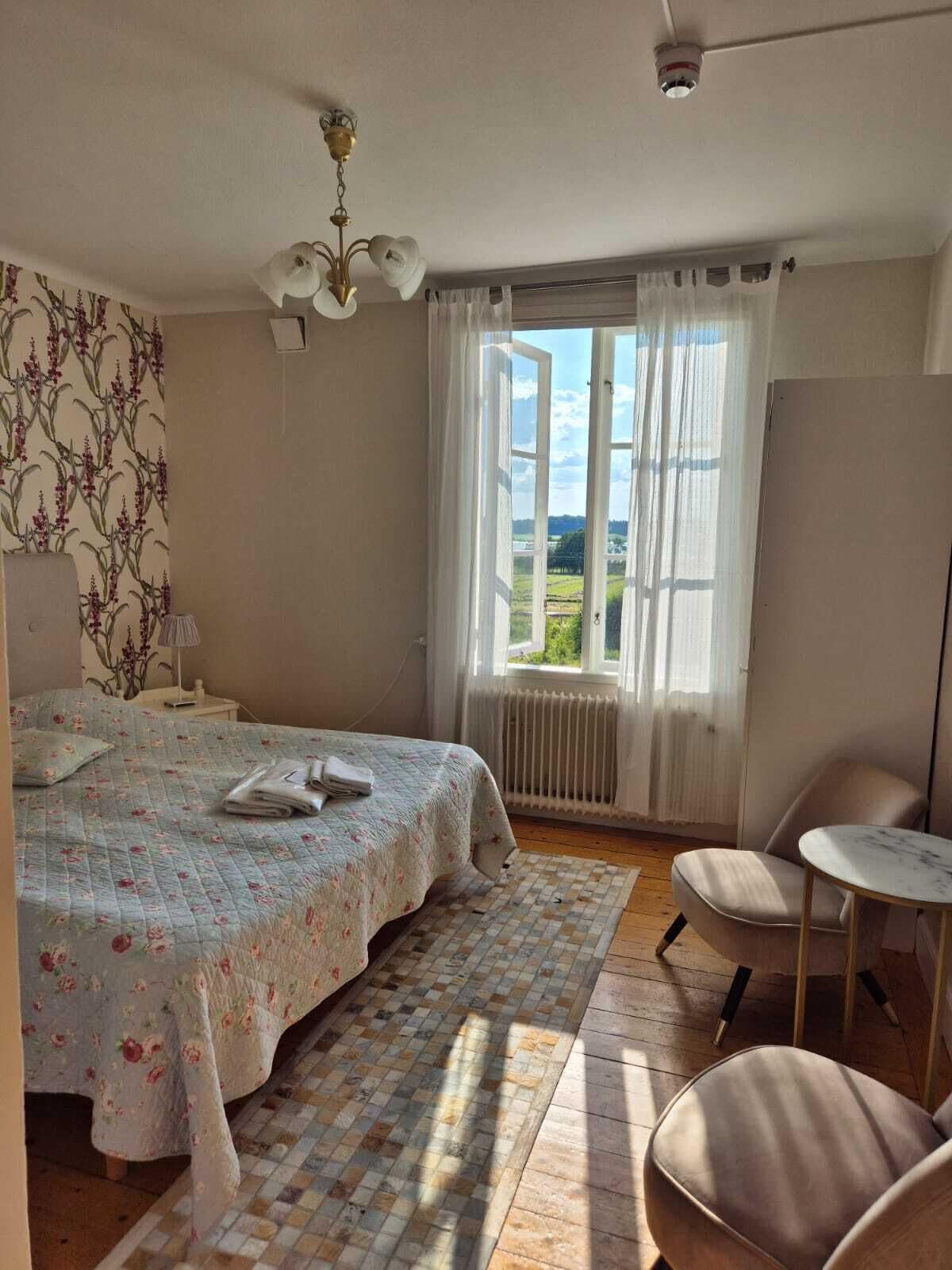 A bedroom with a bed, a chair, a small table, a window with curtains, and a radiator.