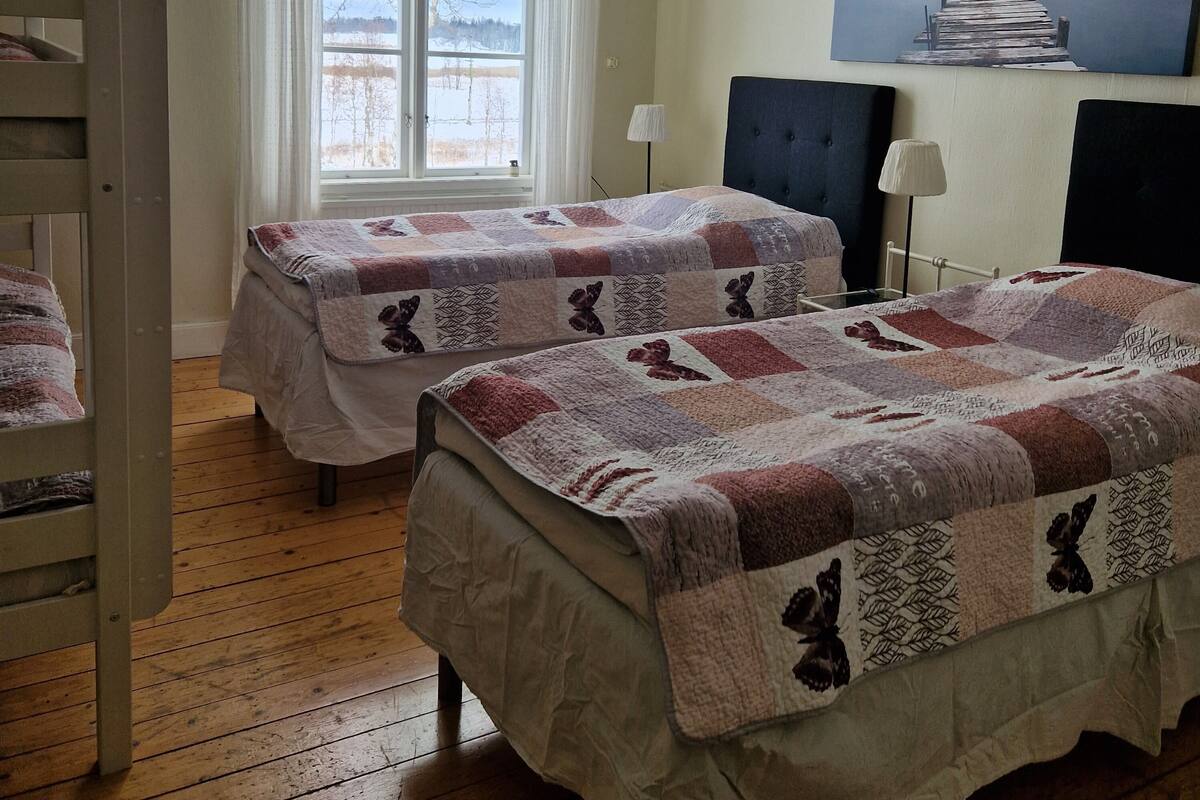 Une chambre avec deux lits, un lit superposé, une fenêtre avec des rideaux, une lampe et un tableau au mur.