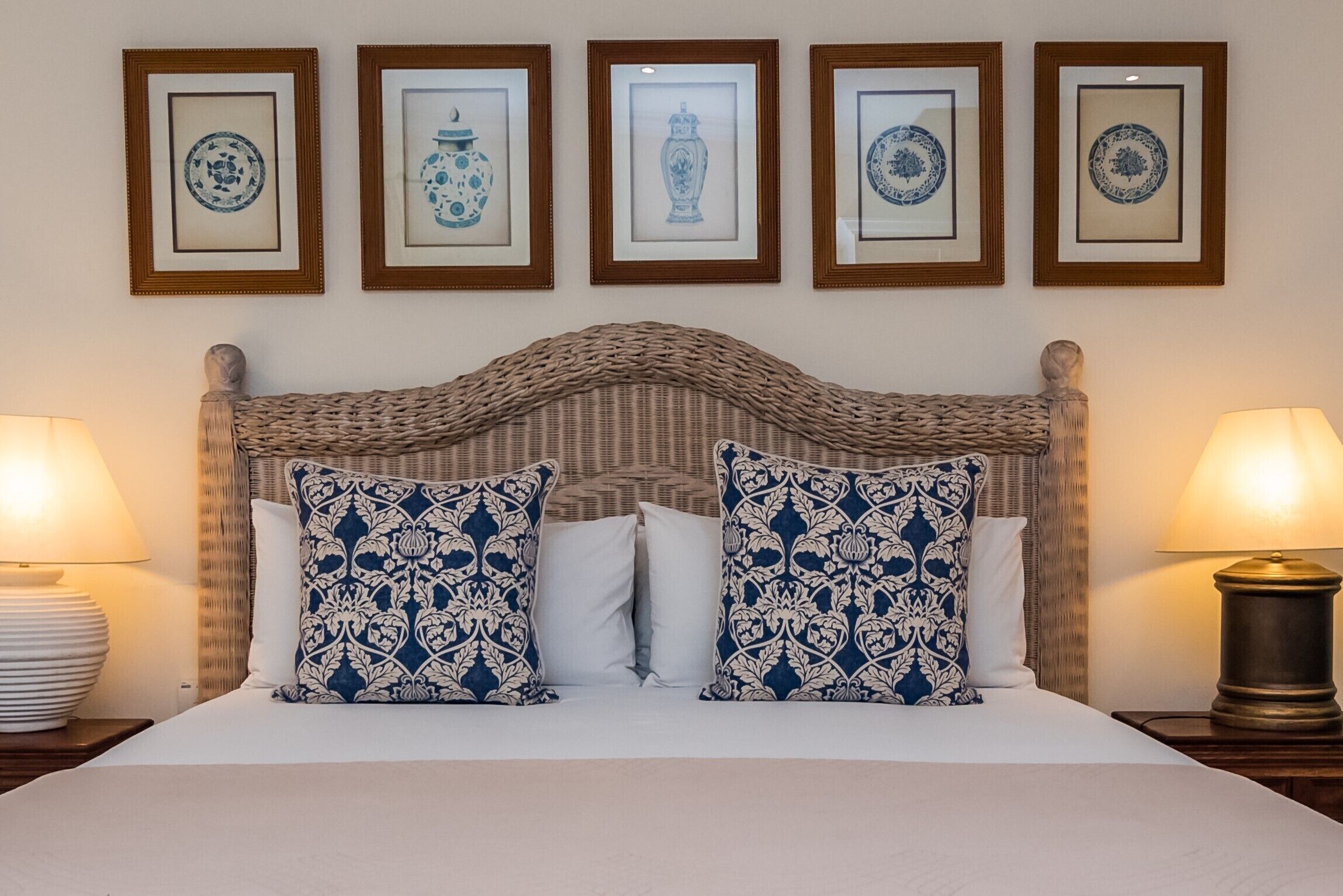 A neatly made bed with patterned pillows, a woven headboard, and framed artwork on the wall.