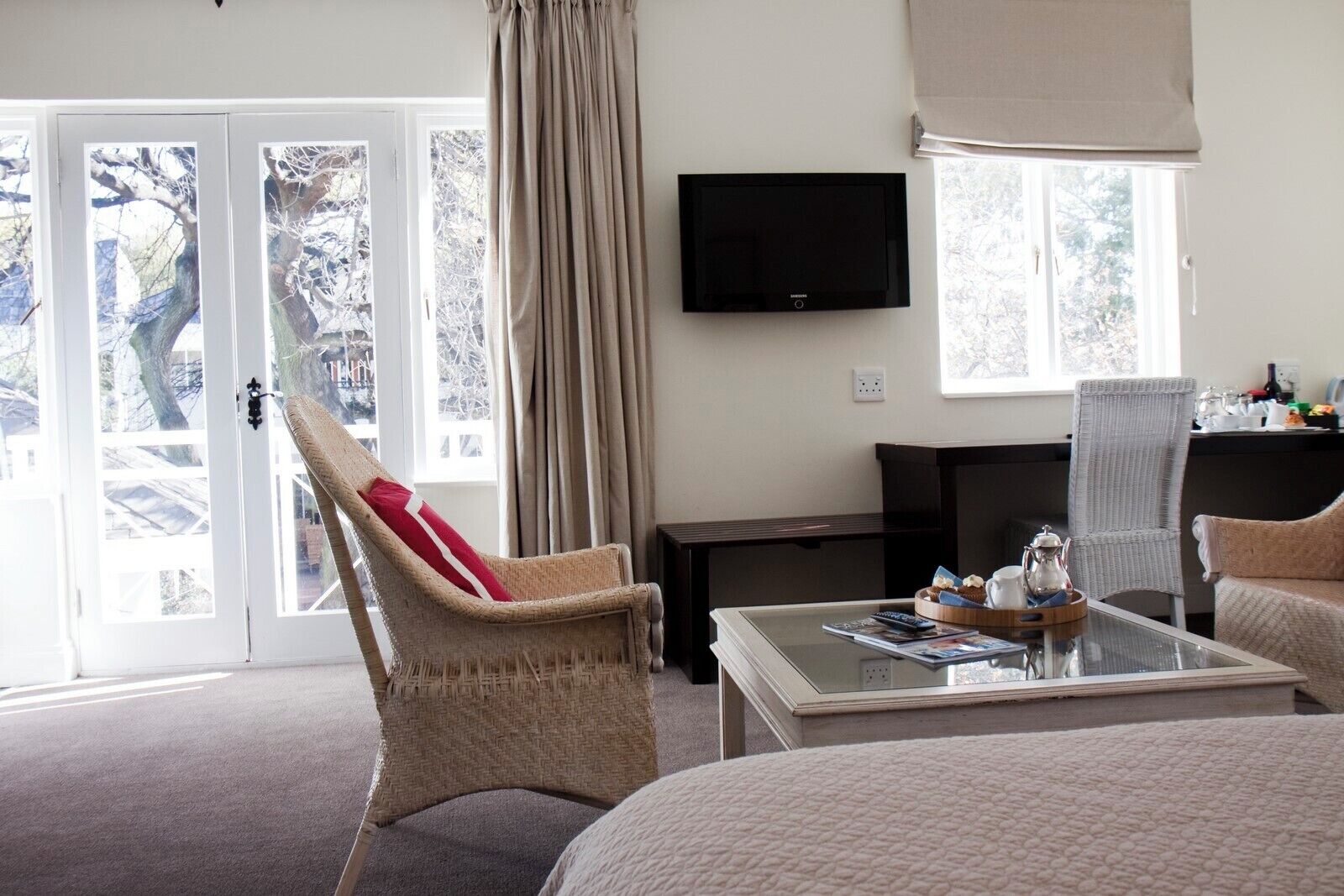 Ein Hotelzimmer mit einem Bett, einem Glastisch, einem Korbsessel, einem an der Wand befestigten Fernseher und Blick auf Bäume durch die Glasüren.