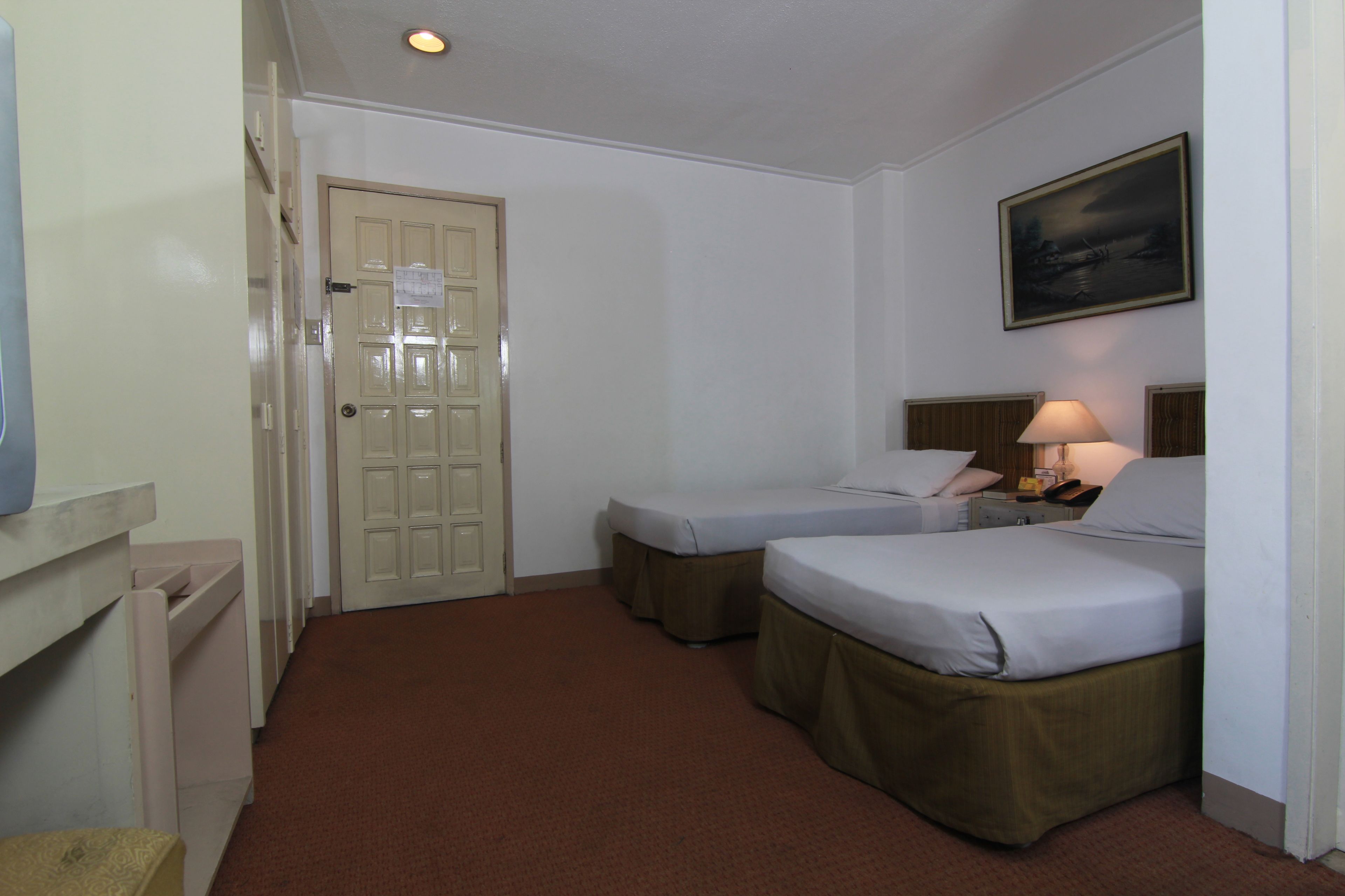 Habitación de hotel con dos camas individuales, una mesita de noche con lámpara, un cuadro en la pared y una puerta con un cartel.
